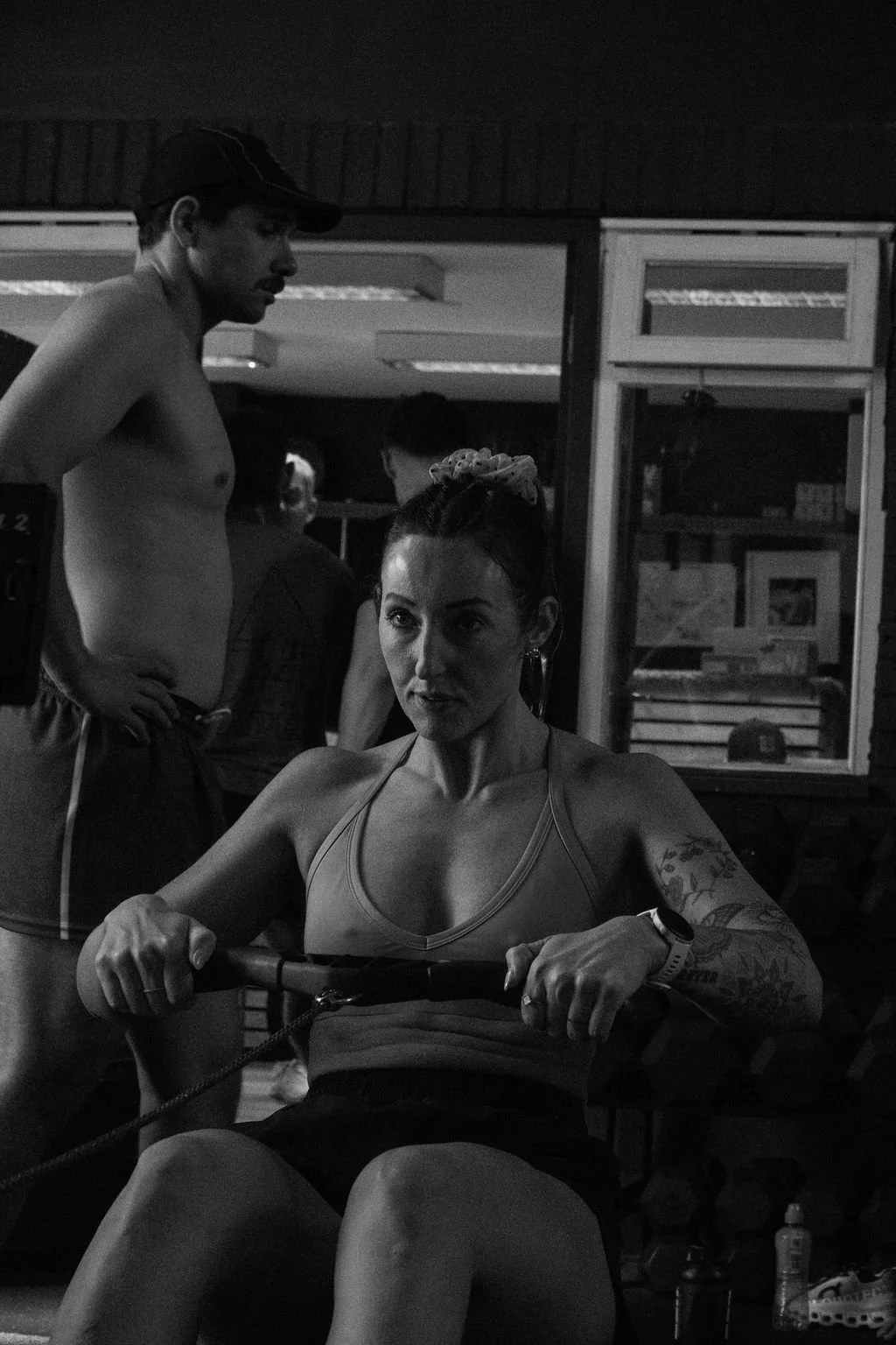 A woman with tattoos and a scrunchie in her hair, sitting on a rowing machine in a gym, while a shirtless man standing beside her looks on.