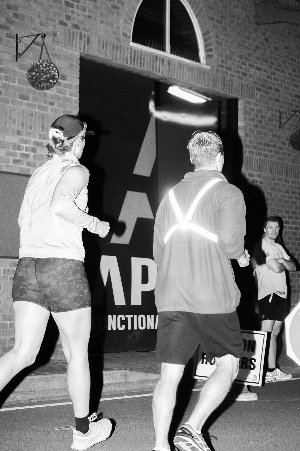 Two runners, a woman and a man, running near a brick wall with a sign and a person standing with crossed arms in the background.