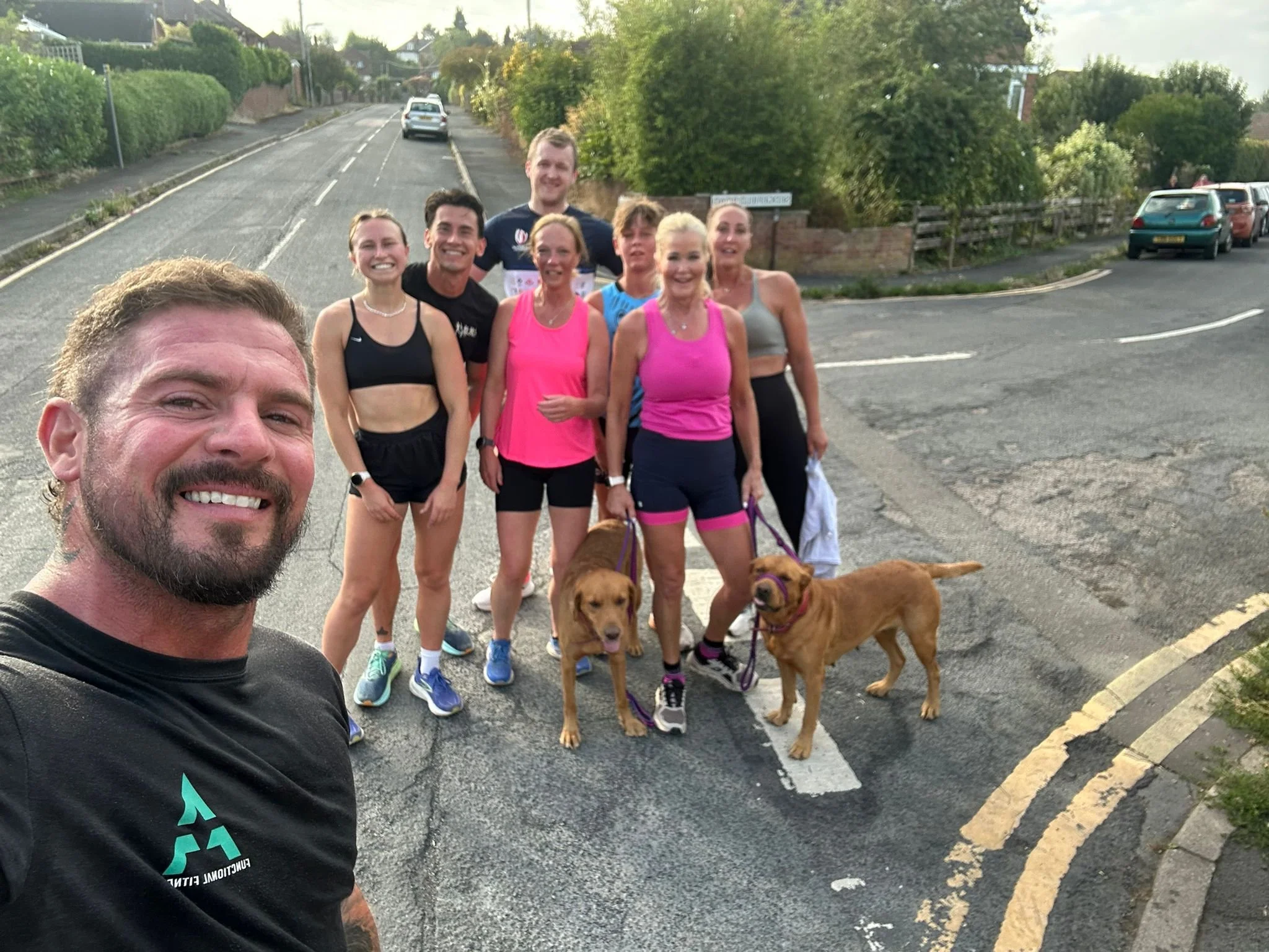 Group of people taking a selfie during a run or walk on a street, with two dogs on leashes, smiling and dressed in athletic clothes.