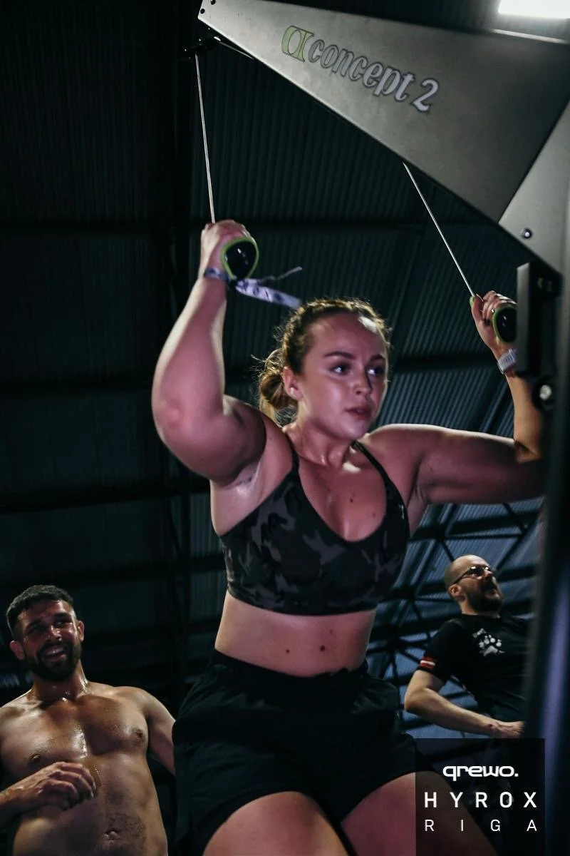 A woman doing a workout using a cable machine at a gym, with two men smiling in the background.