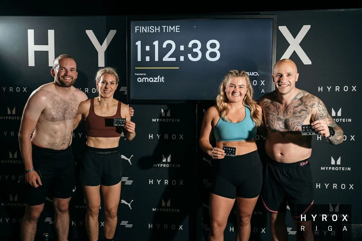 Four people standing in front of a finish line with a digital timer showing 1:12:38, holding Hyrox race tickets, at a fitness event sponsored by Amafit, with a black backdrop featuring logos of Hyrox, MyProtein, Puma, and Amafit.
