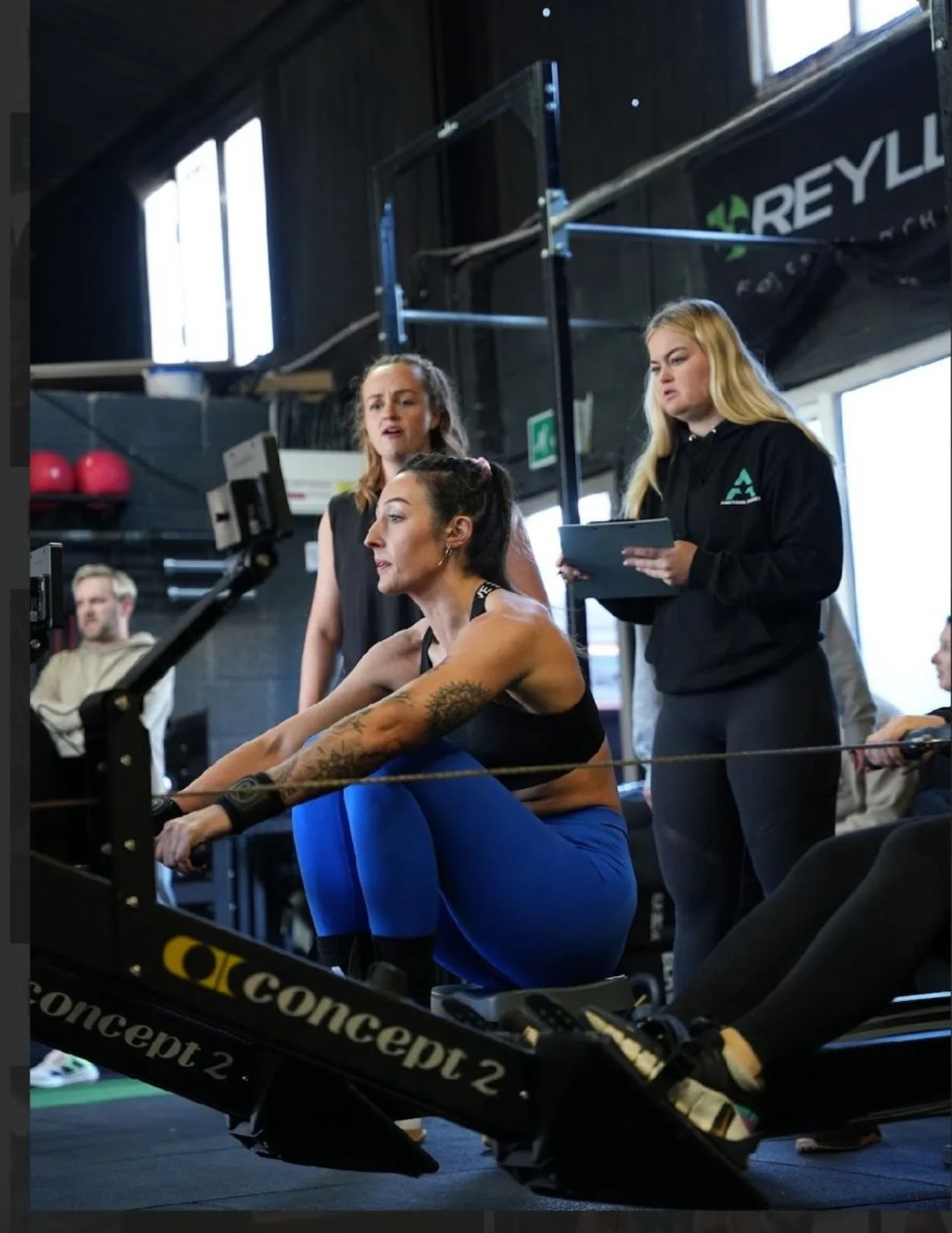 A woman in athletic wear using a rowing machine at a gym, with two women standing nearby observing and taking notes.