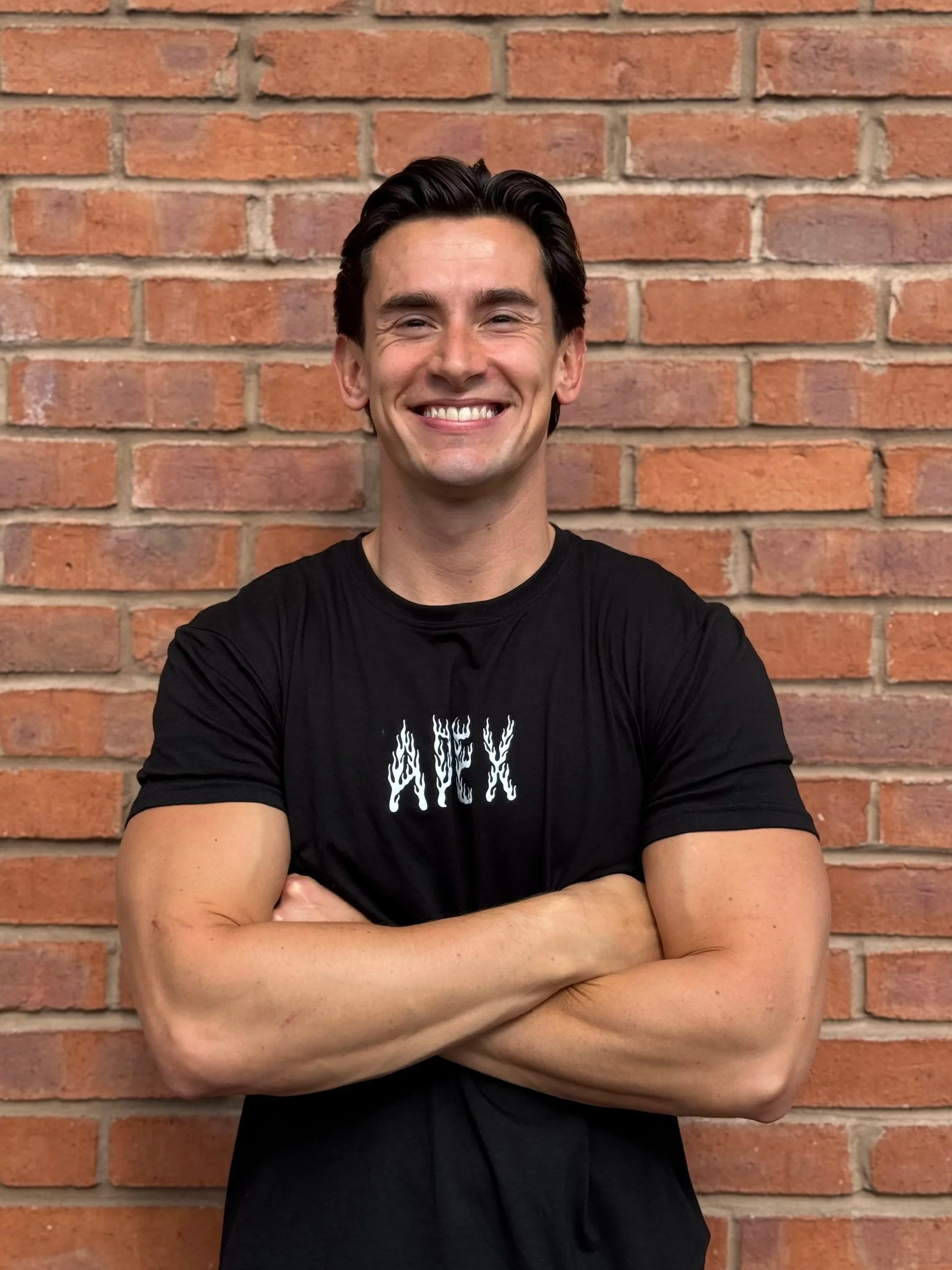 A young man with dark hair smiling and crossing his arms in front of a brick wall, wearing a black T-shirt with the word 'Apex' printed on it.