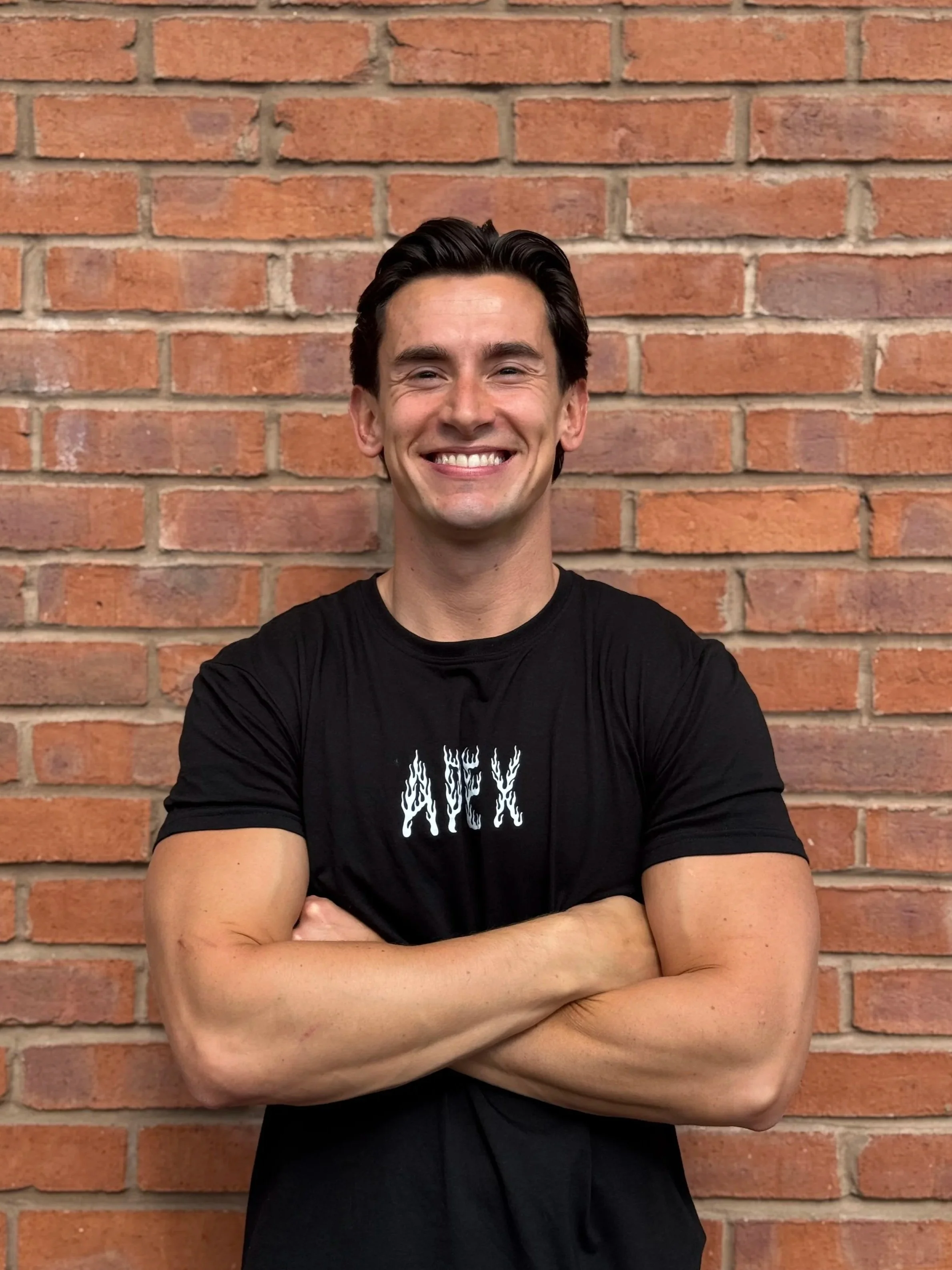 A smiling man with dark hair and fair skin standing in front of a brick wall with his arms crossed, wearing a black T-shirt with white text that appears to say 'WEEK'.