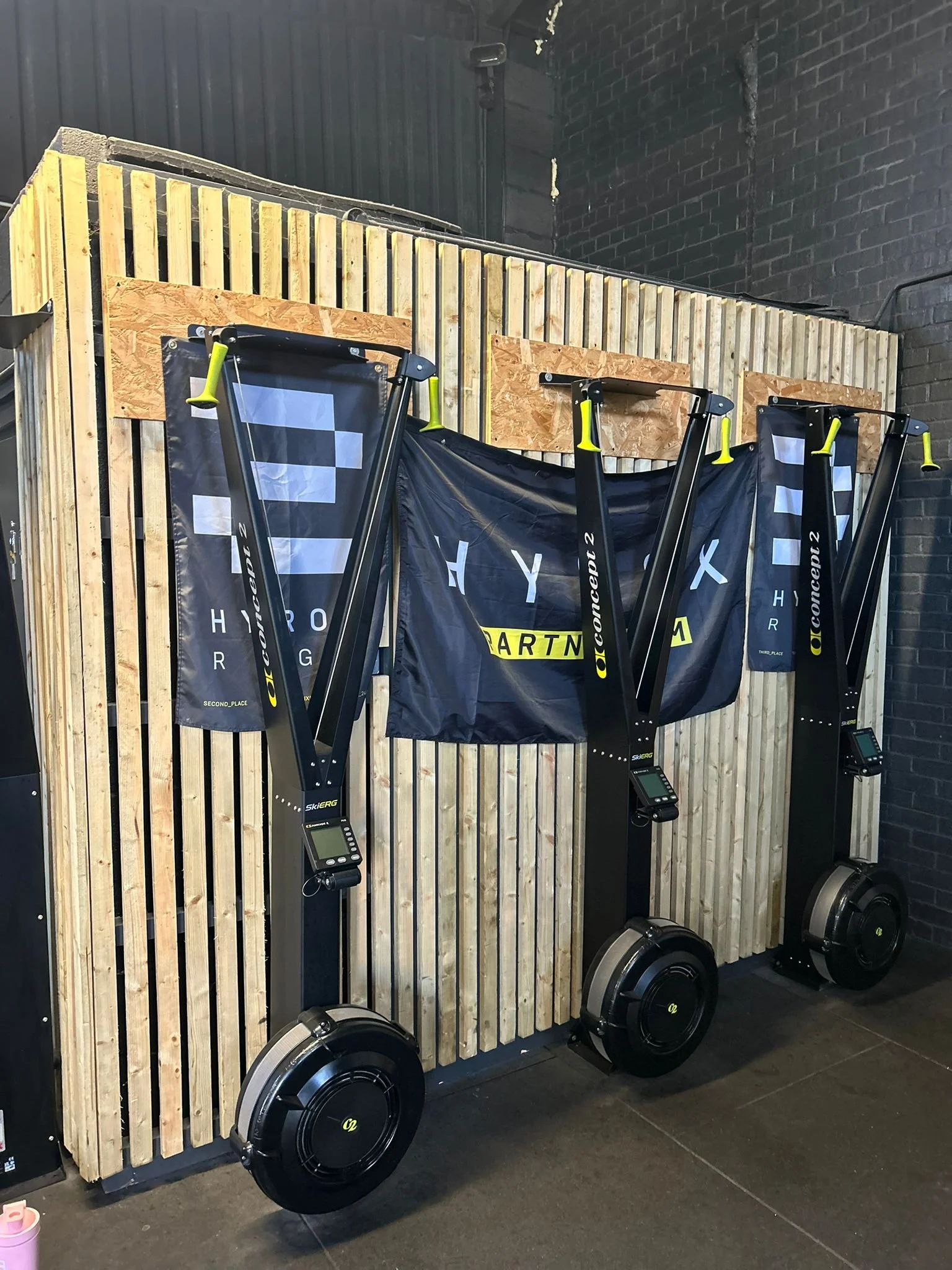 Three black and yellow upright ski-erg machines with digital screens, placed against a wooden slat wall inside a gym.