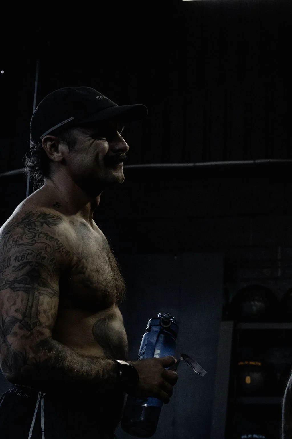A shirtless man with tattoos, wearing a cap, holding a water bottle and a clear cup, standing in a dark gym or training area.