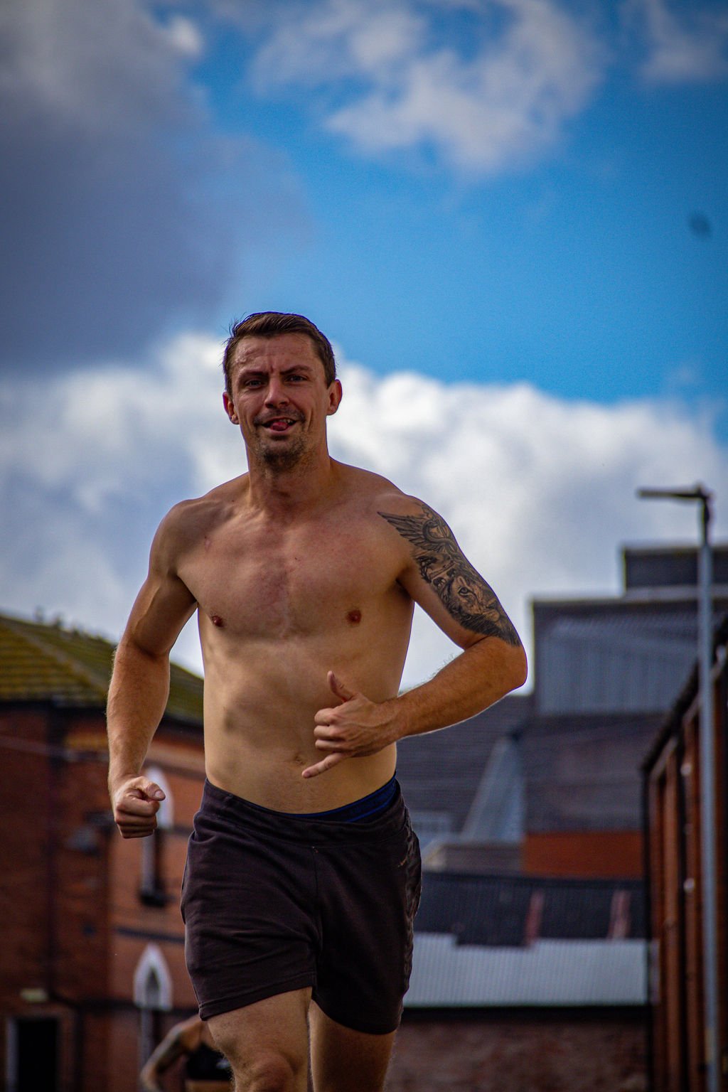 A shirtless man with tattoos running outdoors against a cloudy sky and residential buildings.