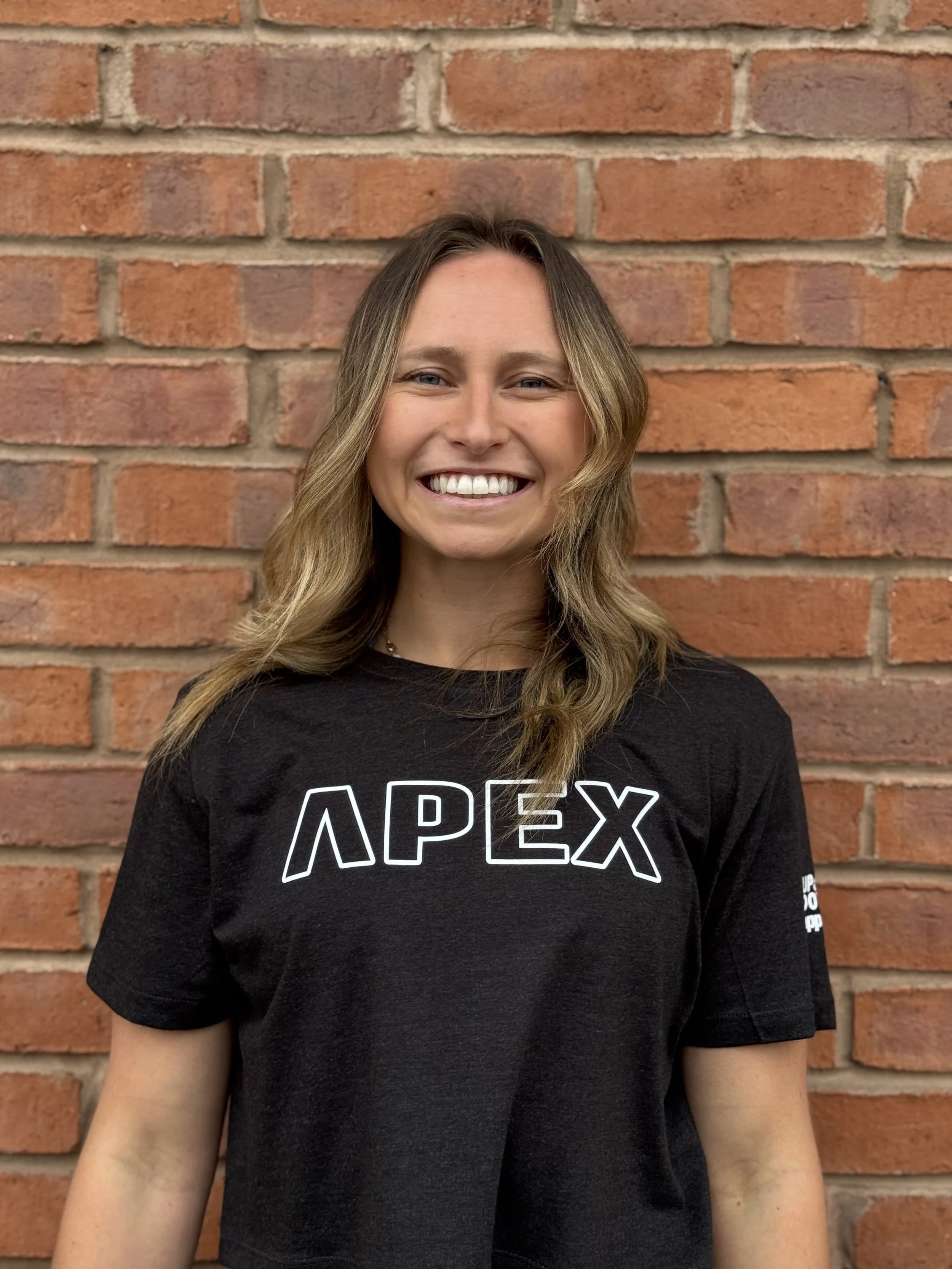 A smiling woman with shoulder-length wavy hair standing in front of a brick wall, wearing a black t-shirt with the word 'APEX' printed on it.