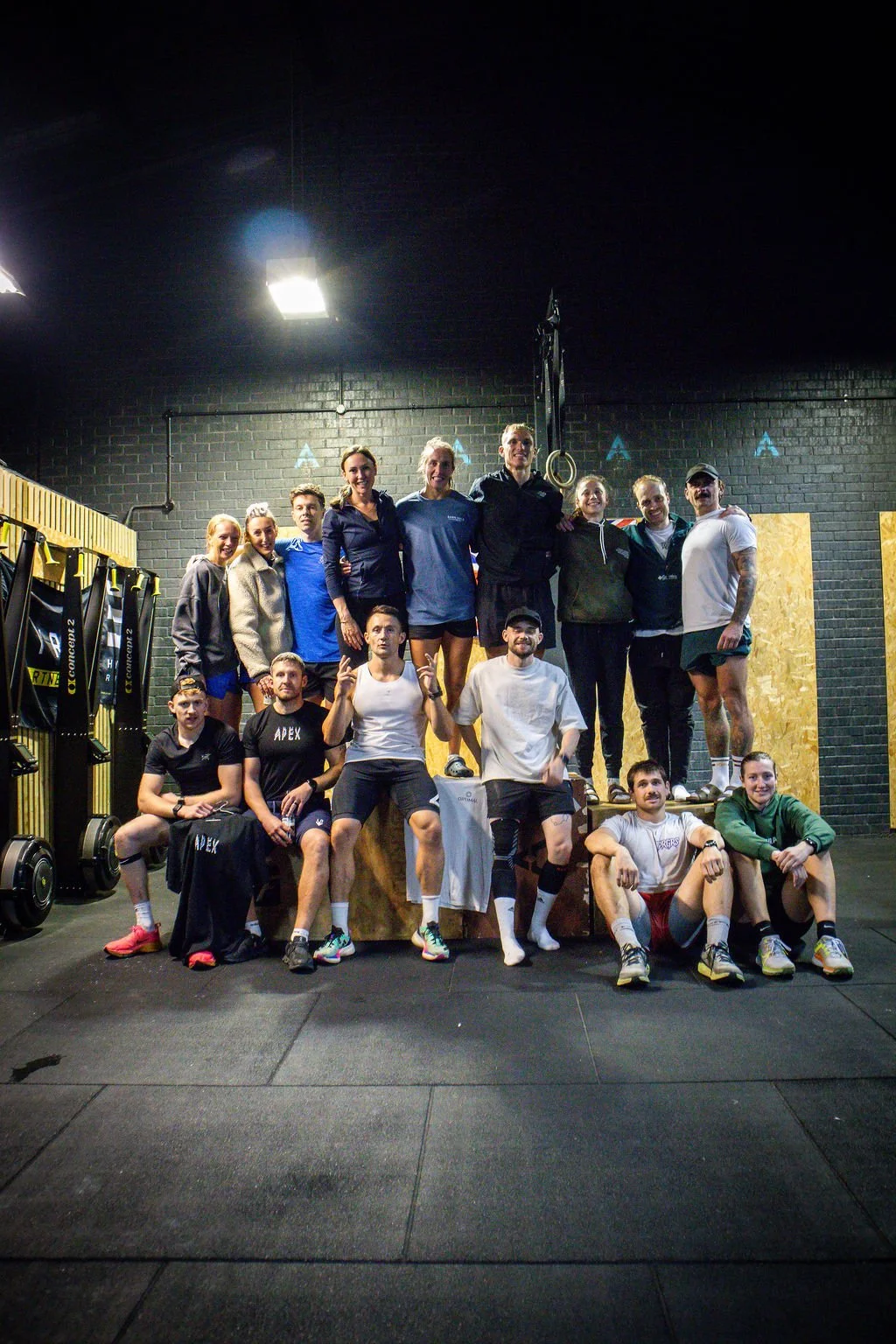 Group of people posing in gym, some standing on a platform, others sitting on the floor, with gym equipment and black brick wall in background.