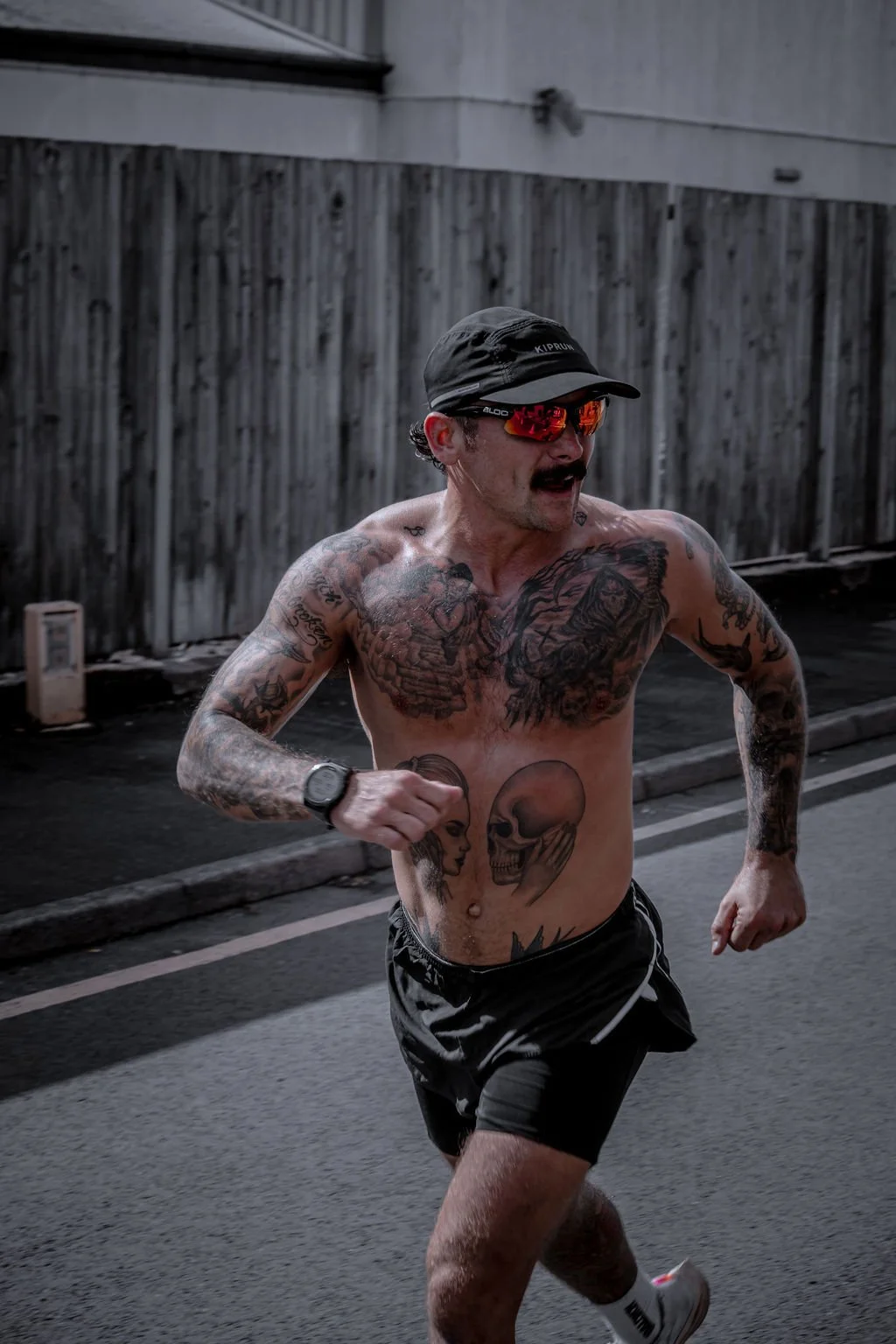 A tattooed man running shirtless on a street, wearing a black cap, sunglasses, black shorts, and a wristwatch.