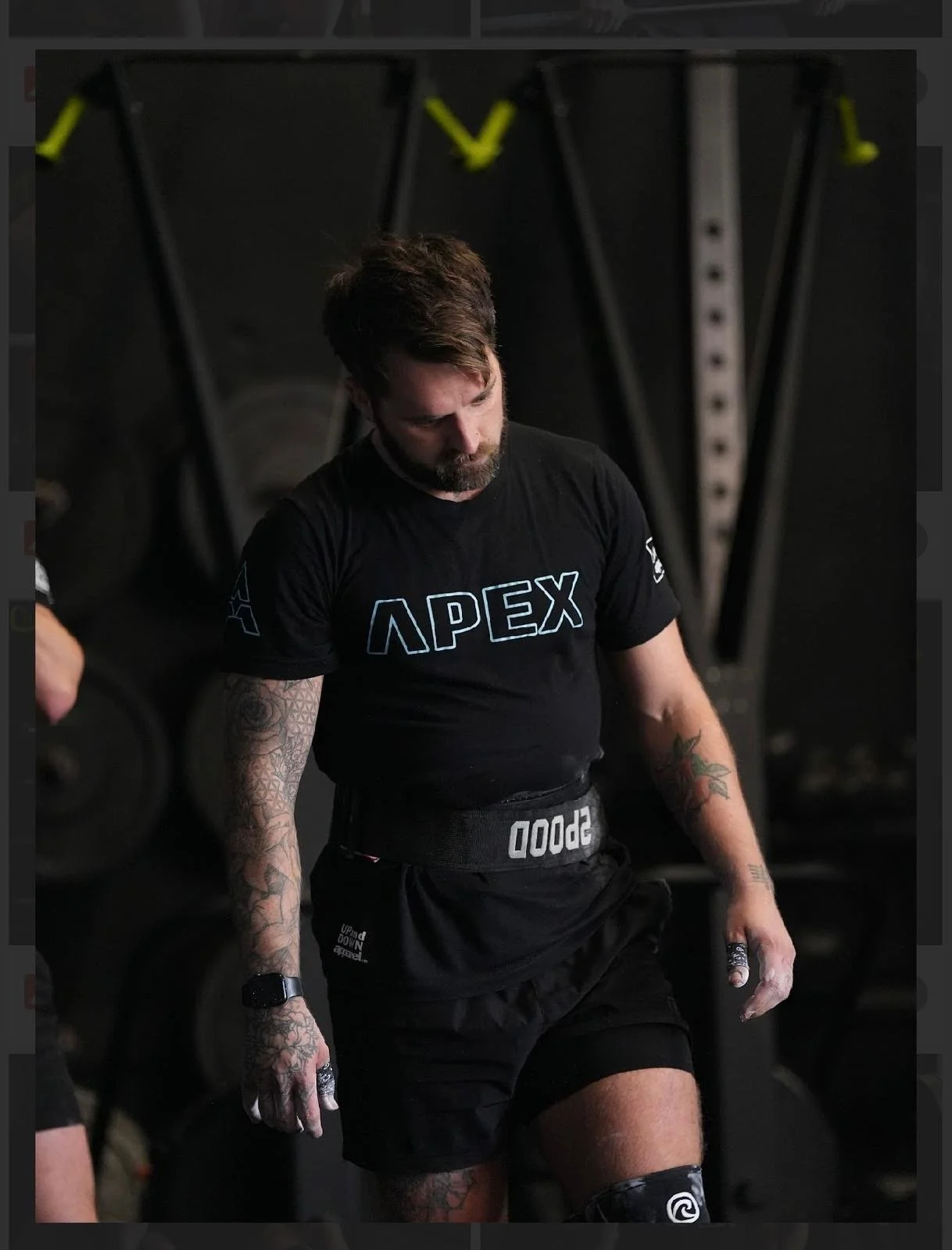 A man with tattoos on his arms, wearing a black Apex shirt, black shorts, and a weightlifting belt, standing in a gym with black equipment in the background.