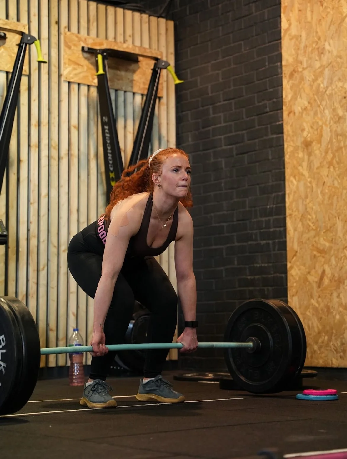 A woman with red hair in a ponytail, wearing a black tank top and black leggings, is deadlifting a barbell with weights in a gym. She is bending forward with her hands gripping the barbell, looking focused.