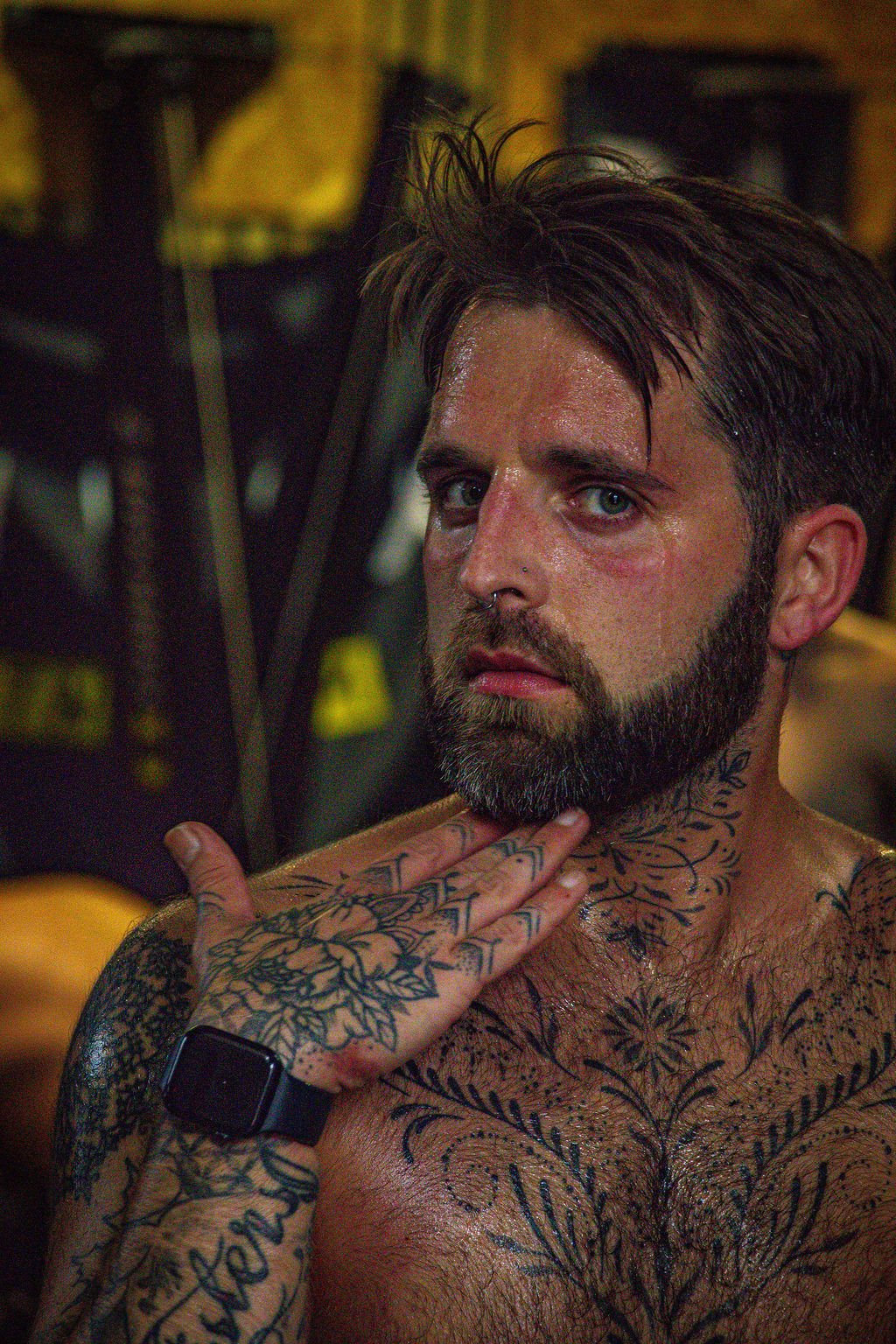 A shirtless man with tattoos on his chest, shoulders, and hand, touching his neck with his hand. He has dark hair, a beard, and is wearing a black wristwatch, with a background of gym equipment.