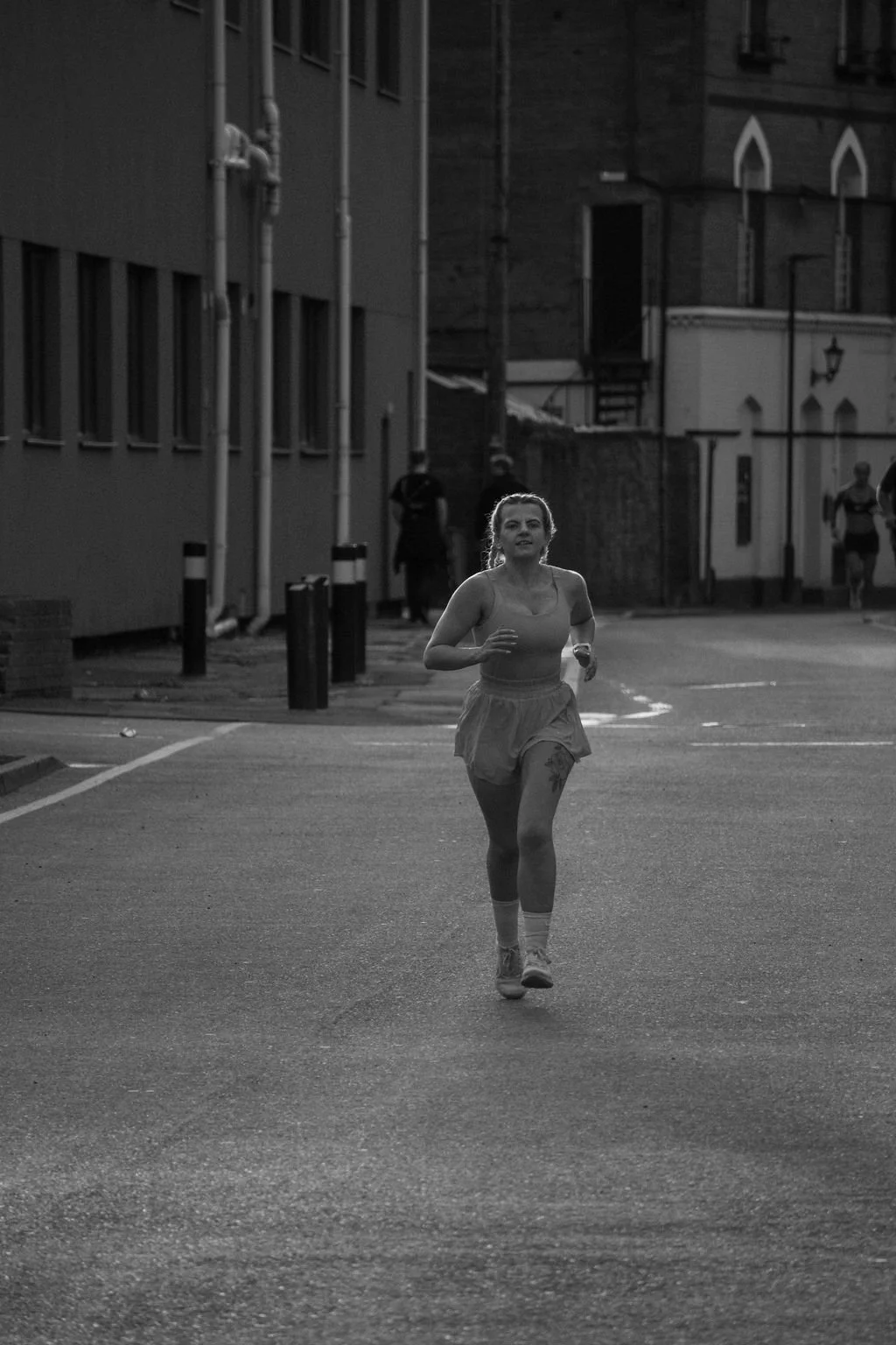 A woman jogging on a city street during dawn or dusk, with a few other runners in the background, in black and white.