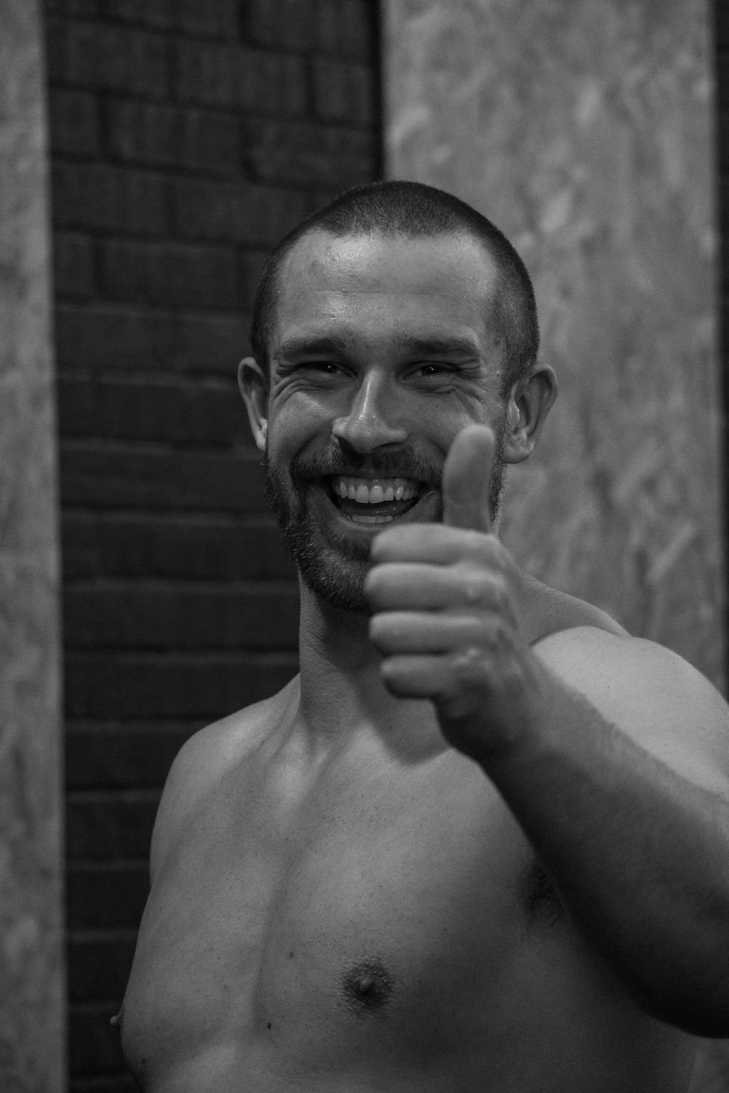 A shirtless man with a beard smiling and giving a thumbs up in front of a background with brick and concrete walls.