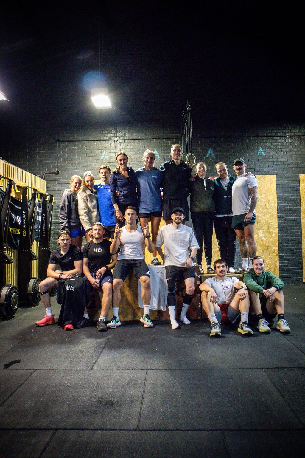 Group of people in a gym, posing for a photo, some standing on a wooden platform, with gym equipment and black brick walls in the background.
