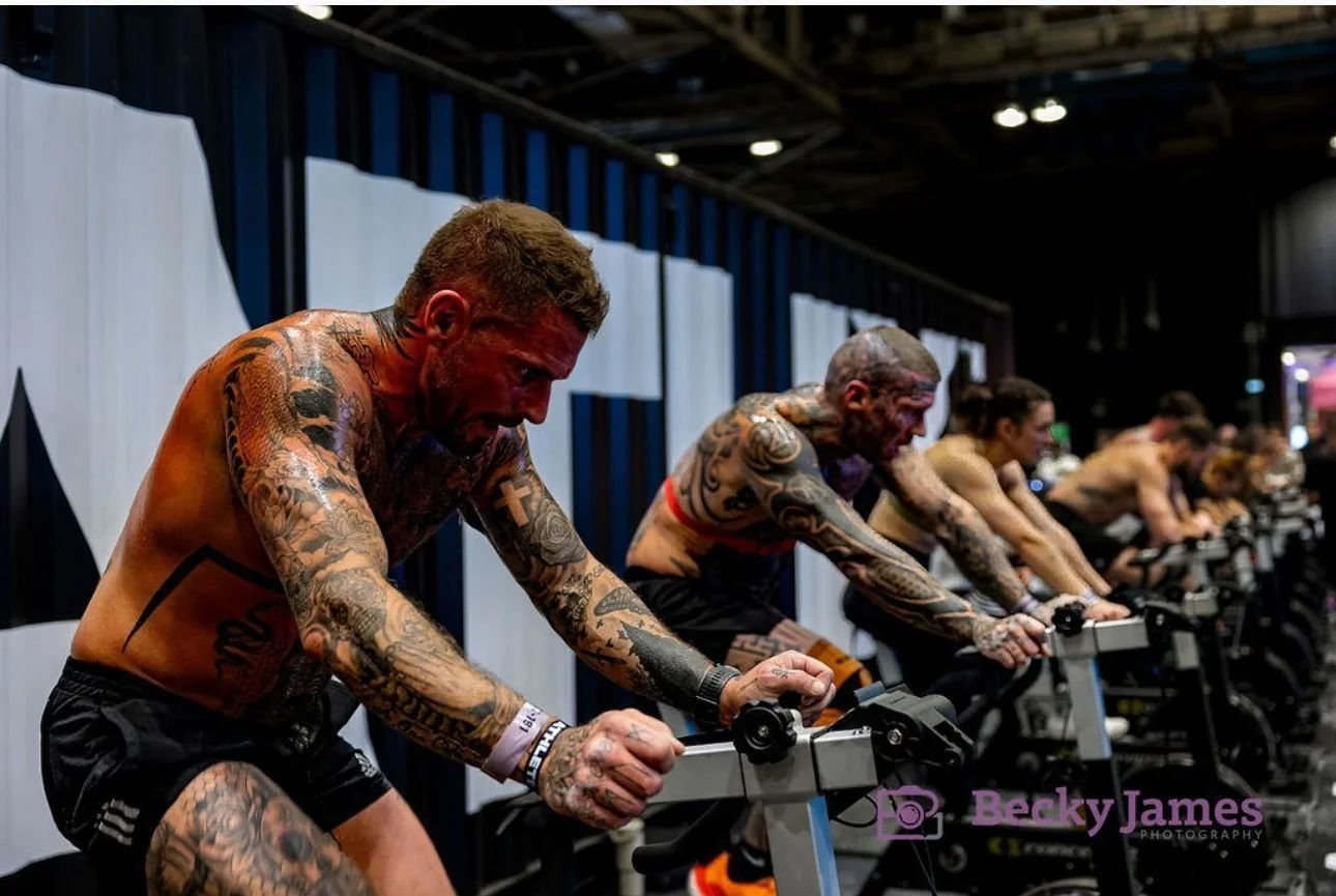 Line of tattooed men with short hair, shirtless and in workout shorts, intensely working out on exercise bikes at an indoor fitness event.