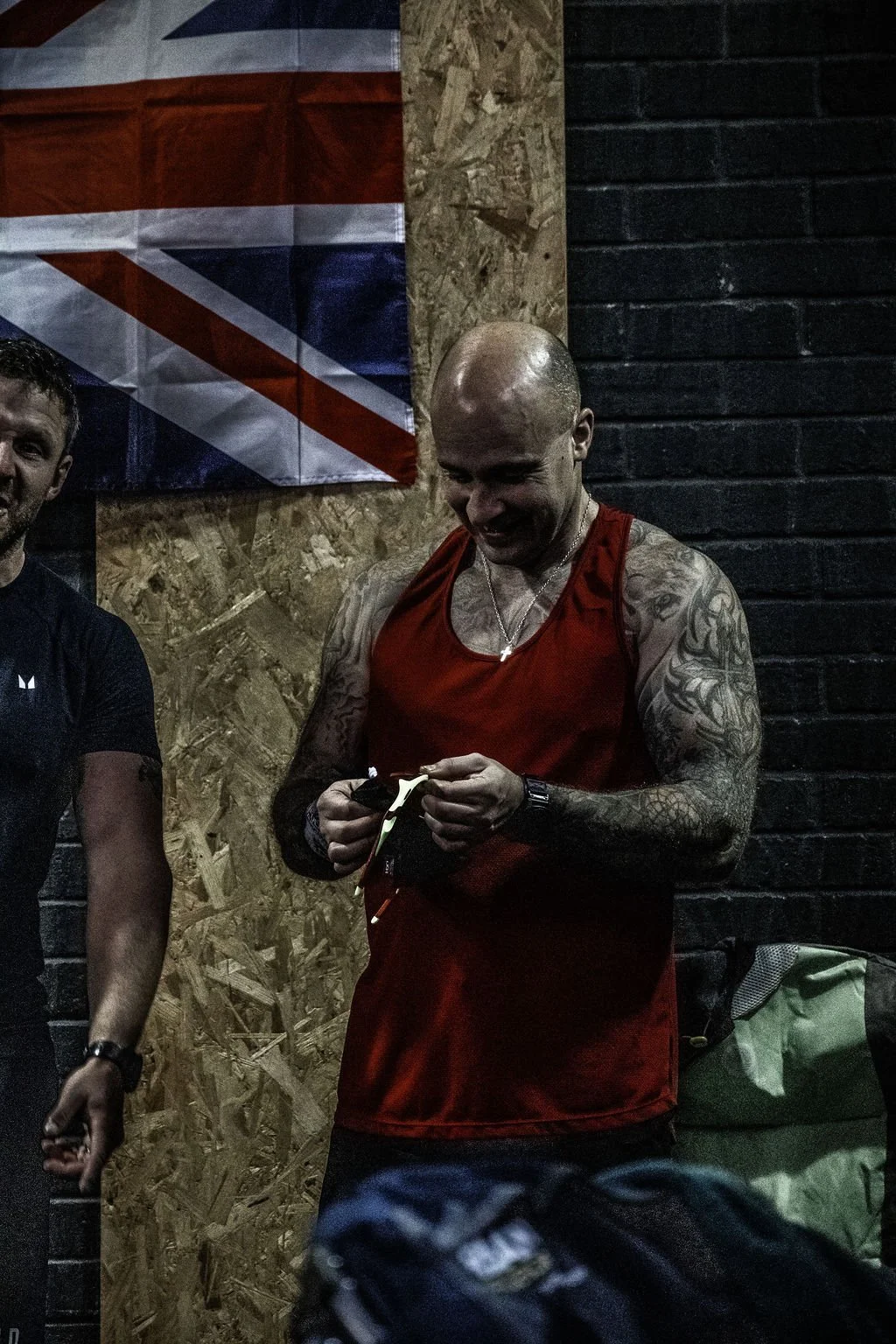 A muscular man with tattoos, bald head, and wearing a red sleeveless shirt is smiling while looking at a small object he is holding. He is standing in front of a black brick wall with a patch of plywood and a British flag hanging behind him.
