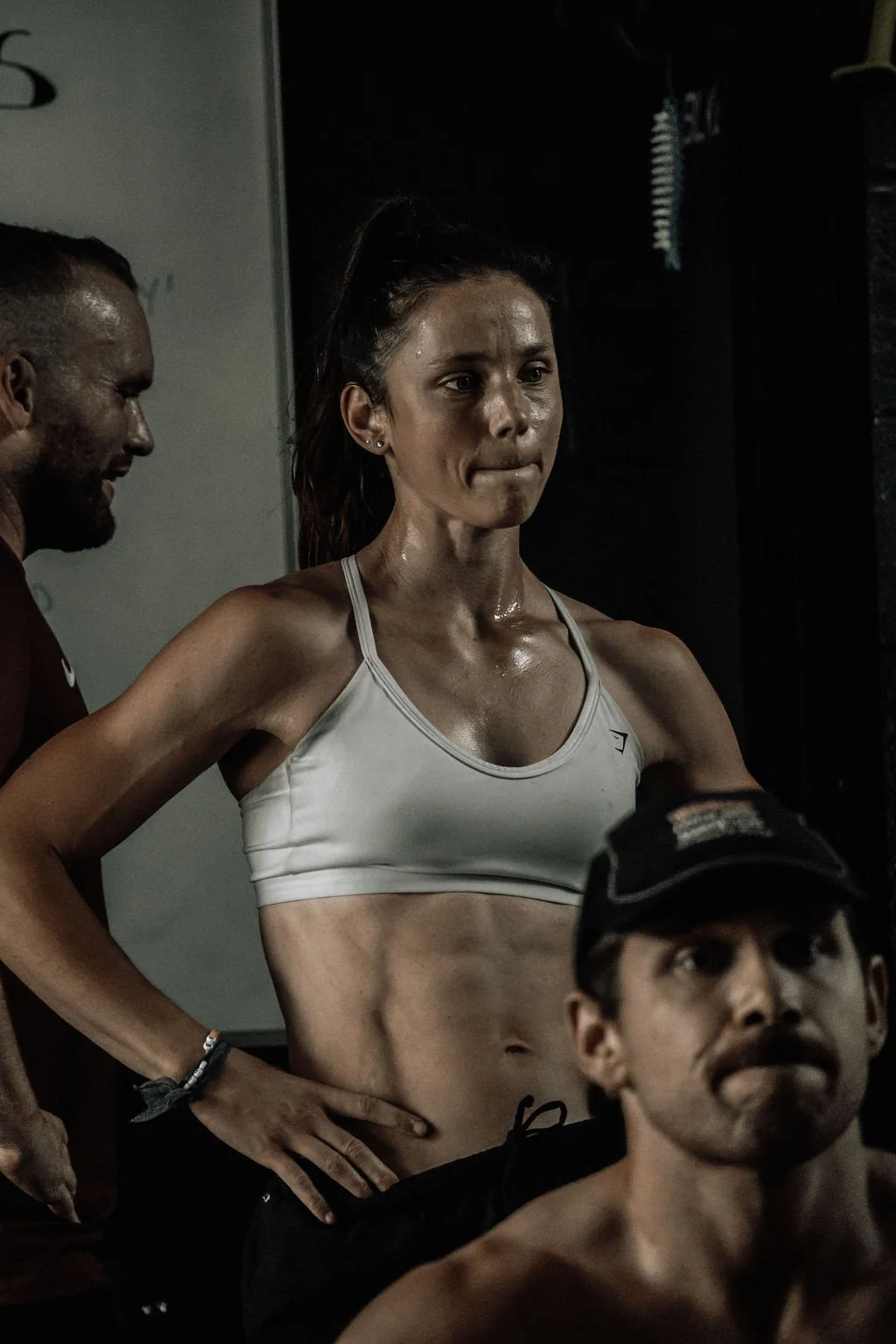 A woman in athletic wear appears to be exhausted or deep in thought, sweating, in a gym or workout setting, with two men nearby, one of whom is partially visible.