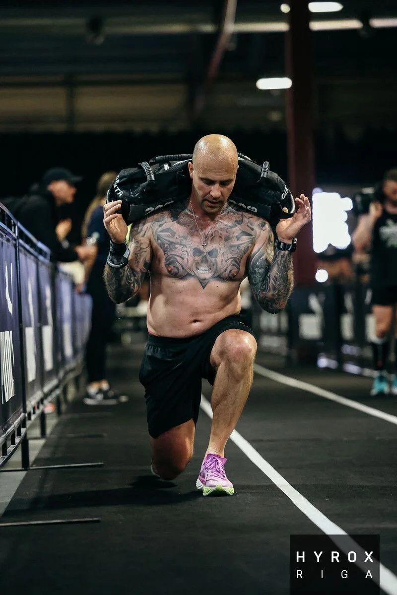 A muscular man with tattoos on his chest and arms, wearing black shorts and purple running shoes, is performing a lunge exercise while holding a weighted sandbag across his shoulders in a gym or training facility with other people in the background.