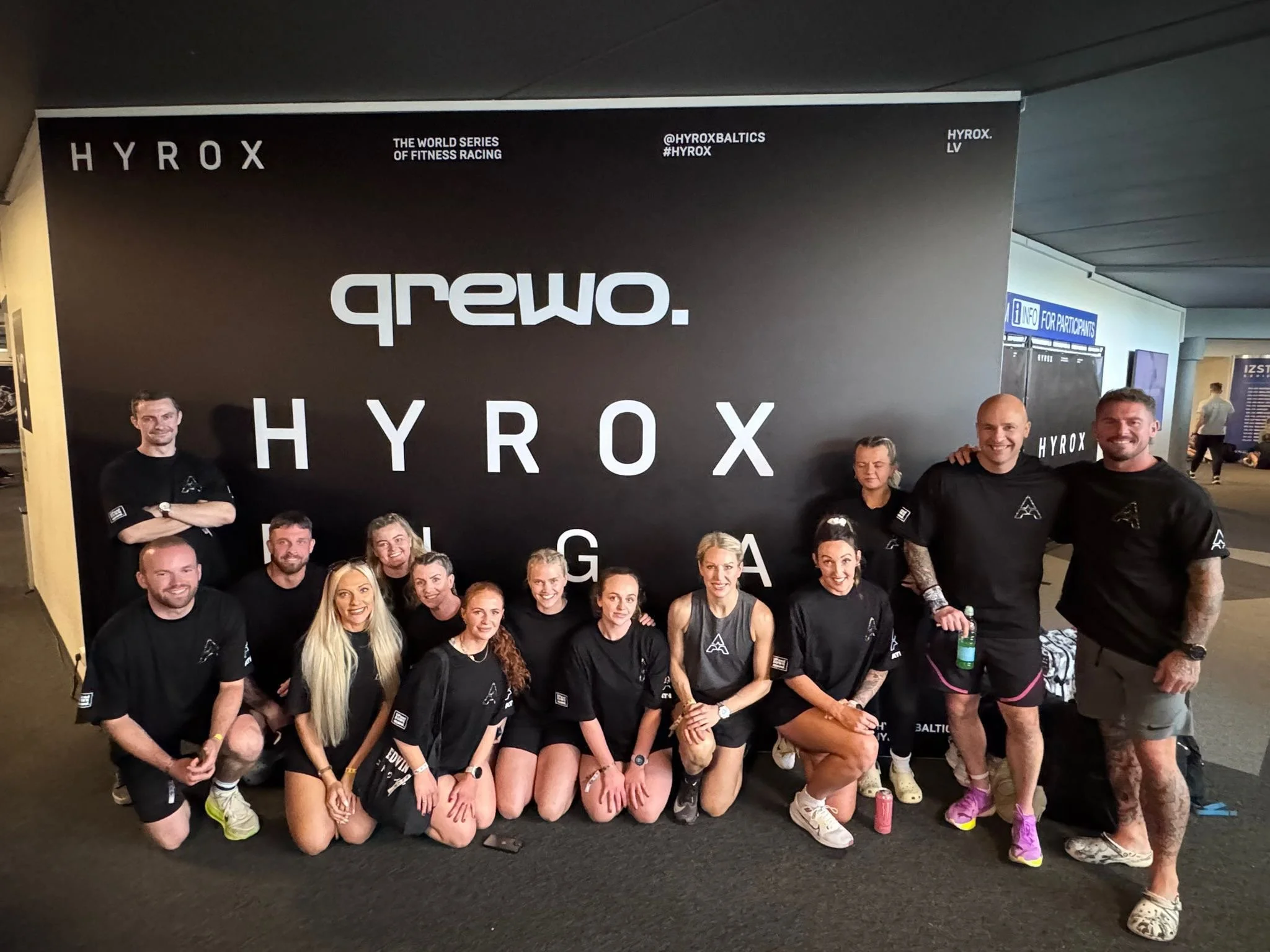 Group of people posing for a photo in front of a large black banner with white text reading 'arewo' and 'HYROX'. The group includes men and women, some standing and some kneeling, wearing athletic clothing.