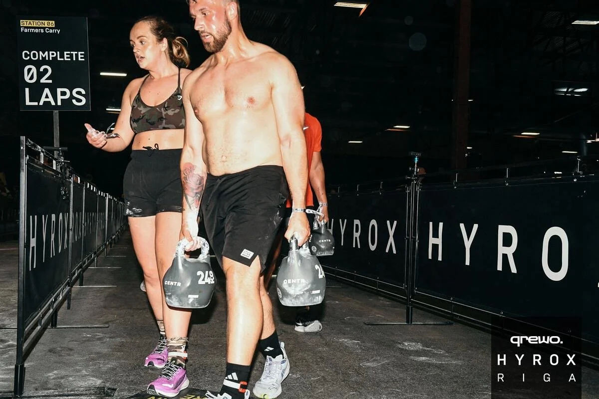 A shirtless man and a woman in athletic clothing carrying kettlebells, walking through a gym or fitness center with branded Hyrox signage, after a race or workout.