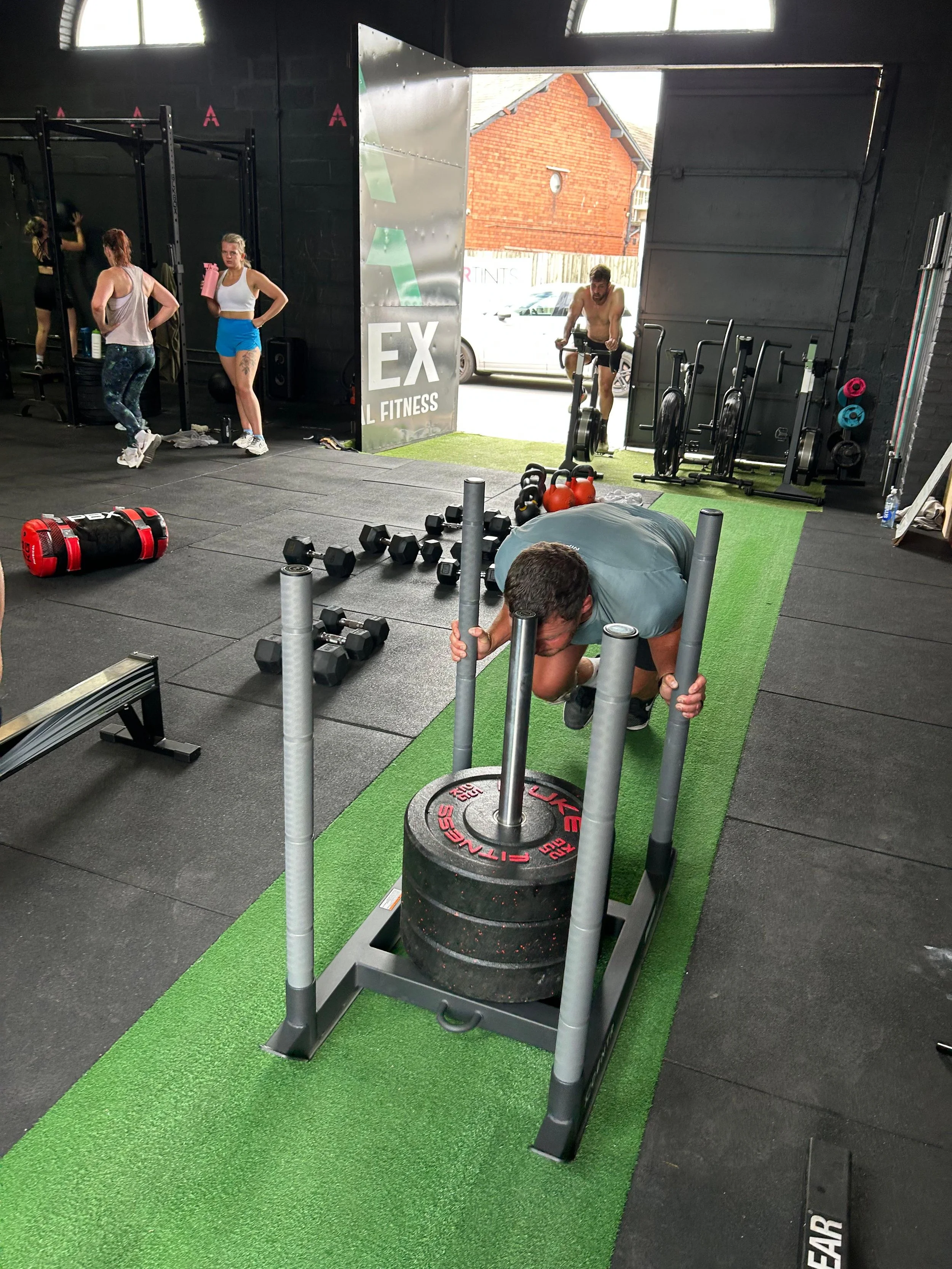 A man pushing a weighted sled in a gym, with various dumbbells, kettlebells, and other exercise equipment scattered around. Two women are standing in the background near a black wall, and a shirtless man is on an exercise bike near the garage door, which is open.