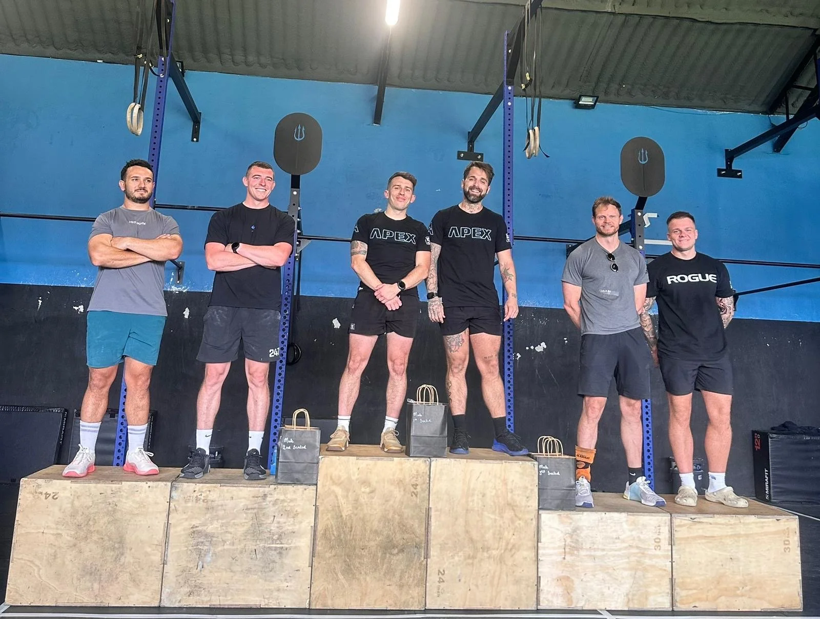Group of six men standing on podium blocks inside a gym, with fitness equipment like rings and pull-up bars in the background, smiling and posing for a photo.
