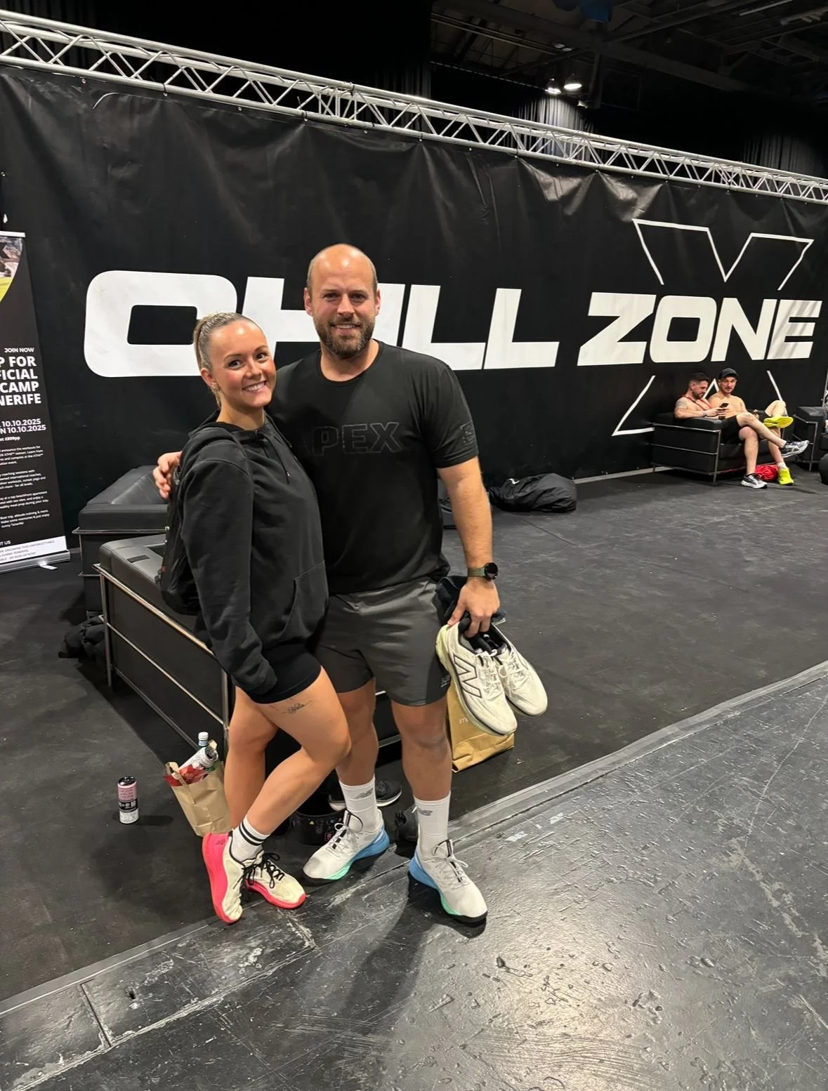 Two people standing together in a gym, smiling at the camera. The woman is wearing a black hoodie, black shorts, and sneakers, while the man is dressed in a black T-shirt, gray shorts, and holding a pair of sneakers. In the background, there is a lar