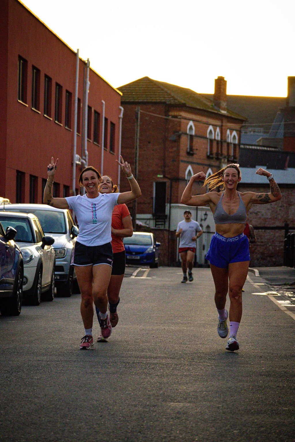 Five women running on a city street during sunset, smiling and making victory signs, with parked cars on one side and brick buildings in the background.
