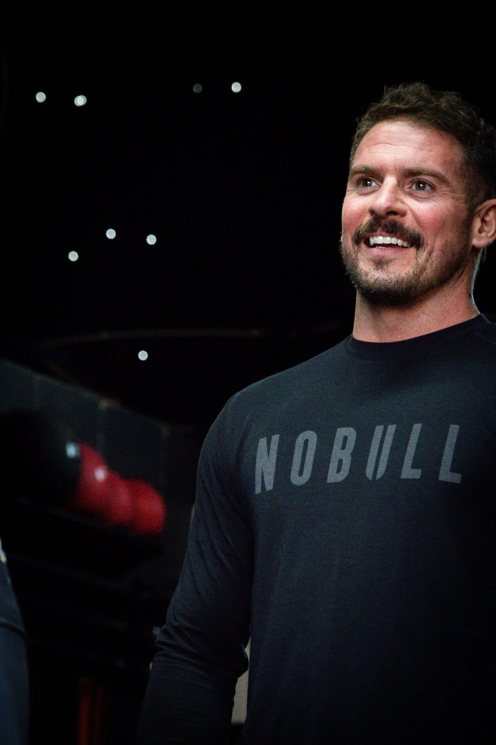 A smiling man with a beard wearing a dark gray shirt with the word 'NOBULL' on it, standing indoors with a dark background and small lights overhead.