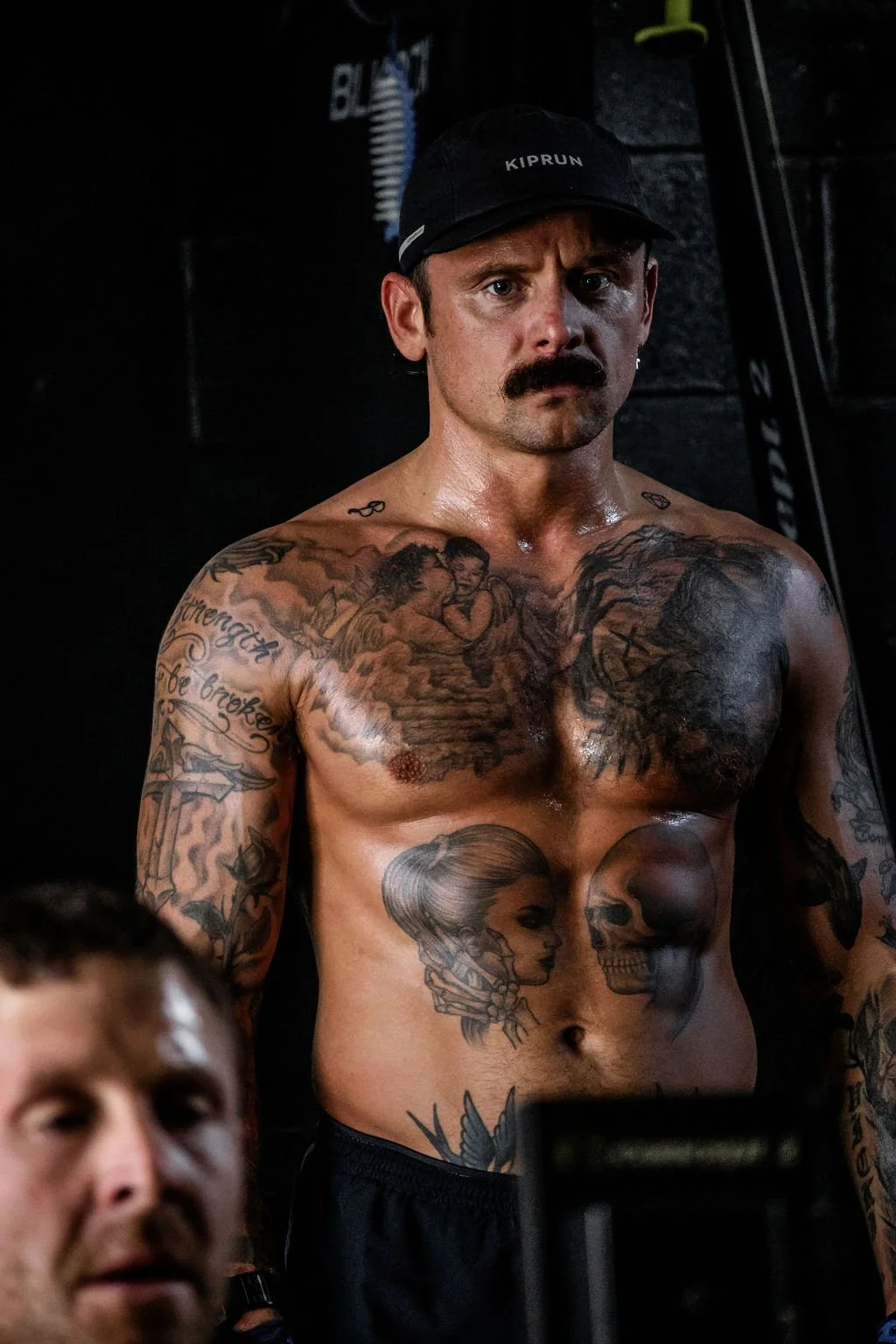 A shirtless man with tattoos, a thick mustache, and a black Kiprun cap looking directly at the camera in a gym setting.