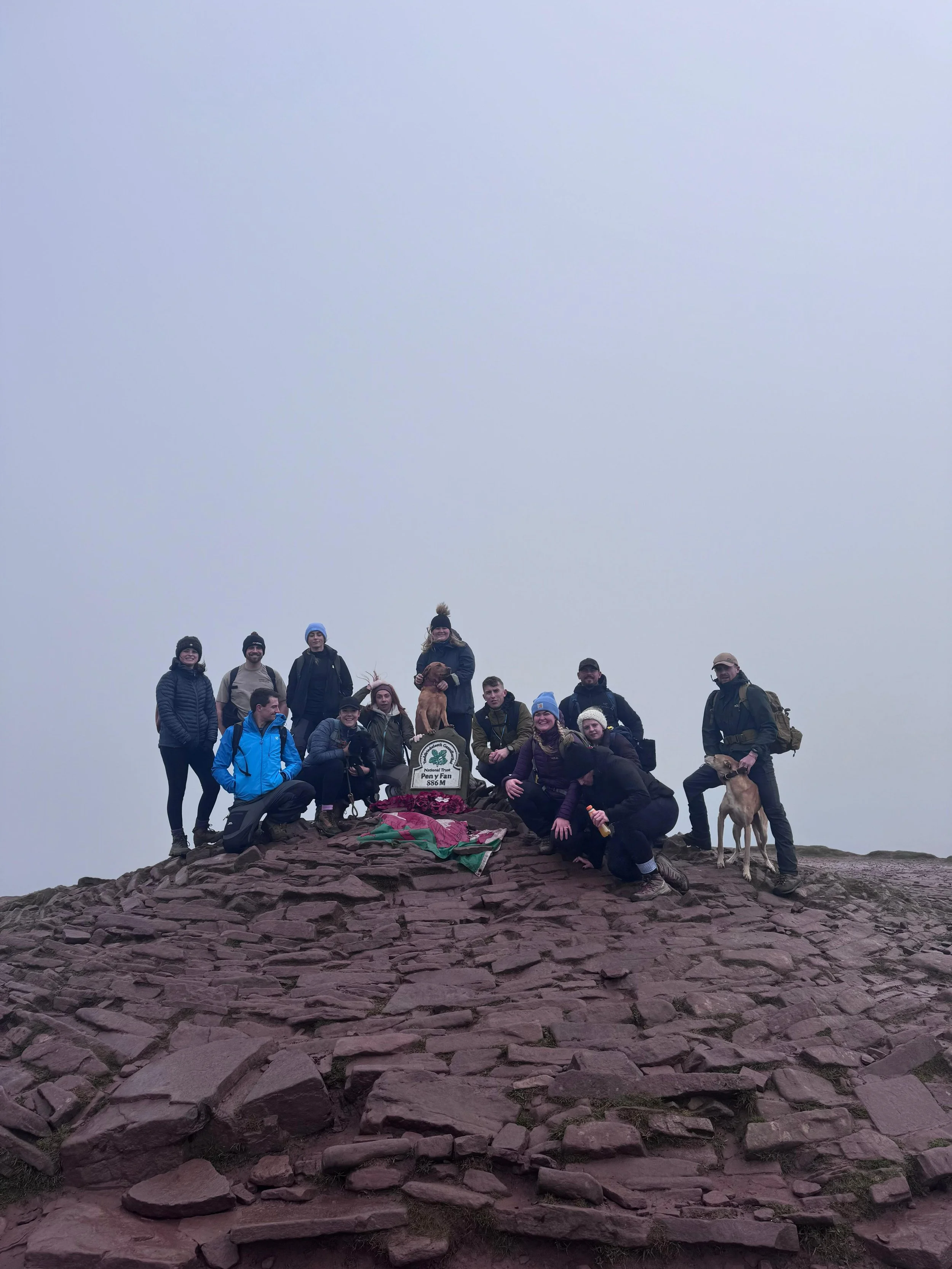 Group of hikers on a rocky mountain summit with a sign that reads 'Pen y Fan 886 m' and dogs, under a gray overcast sky.