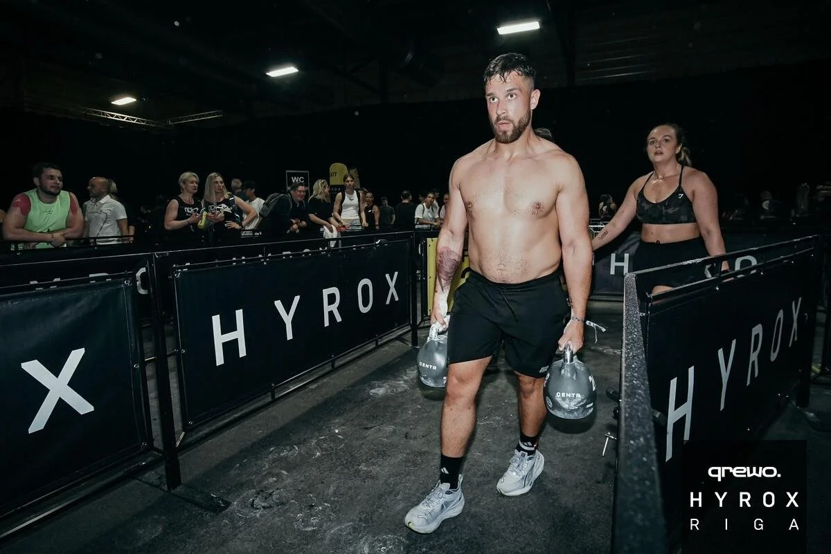 A shirtless man with a beard and tattoos on his left arm walking through a fitness competition arena holding two water bottles, with a woman walking behind him and other spectators nearby, at the HYROX Riga event.