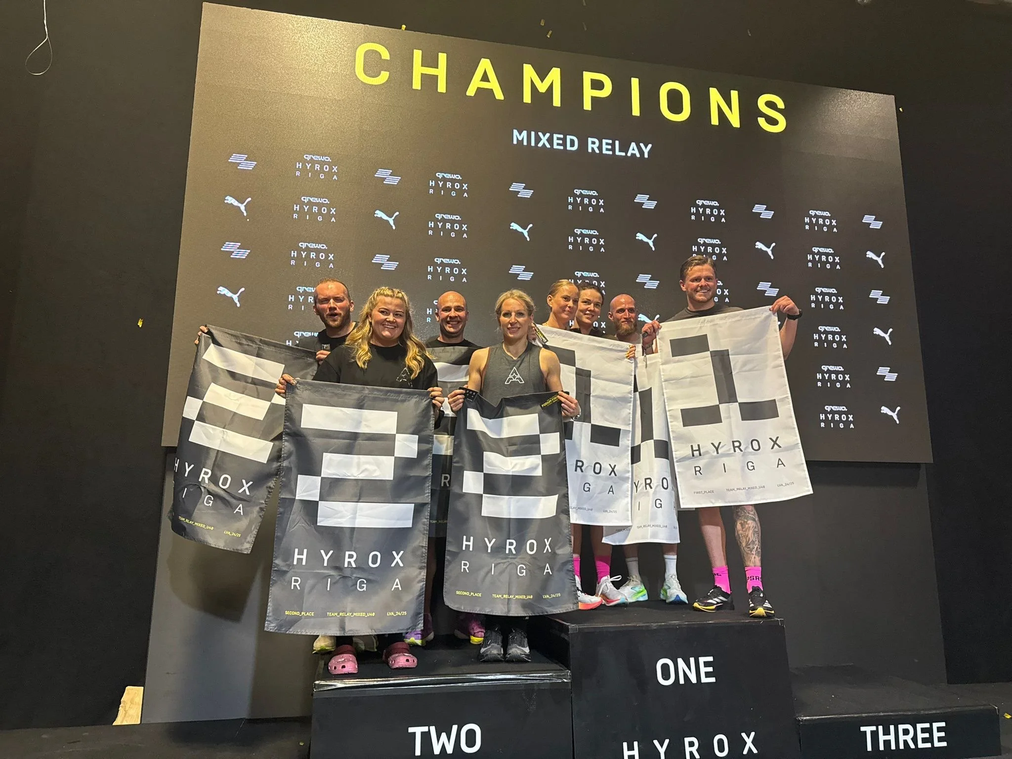 Group of seven people on a winners' podium celebrating after a relay race at the Hyrox Riga event, holding flags with the Hyrox logo, with a large screen behind them displaying the word 'CHAMPIONS' and other text.
