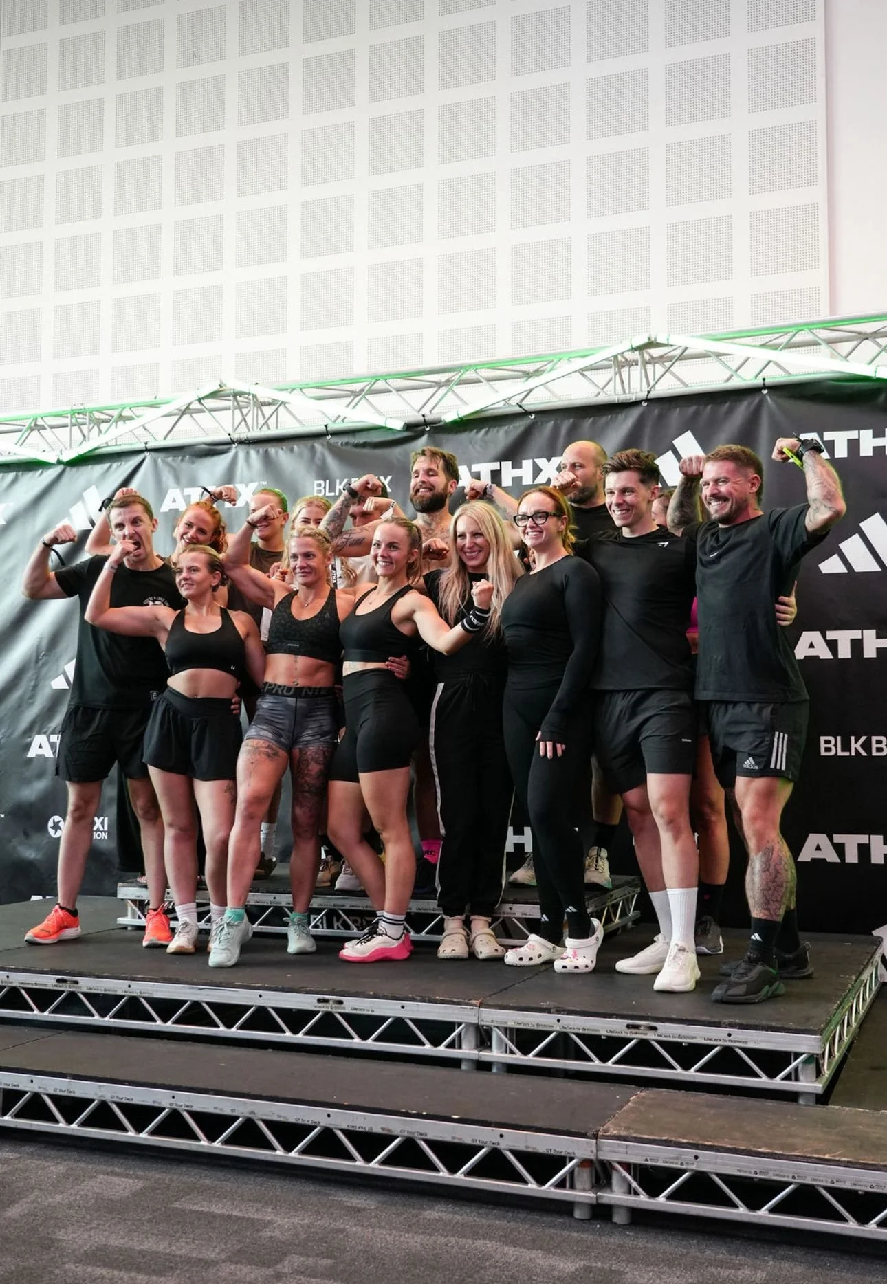 Group of athletic men and women posing with flexed arms on a stage, at a fitness event, in front of a black backdrop with white logos.