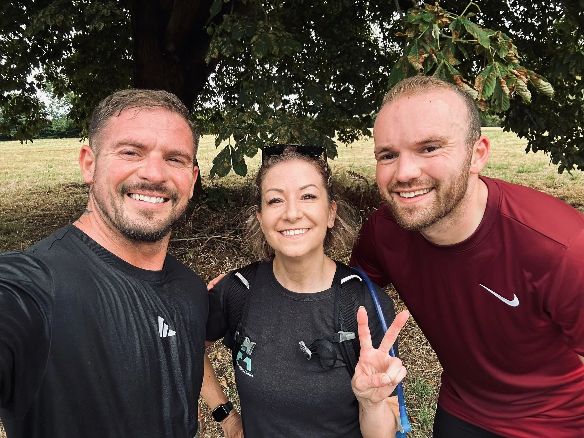 Three smiling people taking a selfie outdoors under a tree, with one woman flashing a peace sign, all wearing athletic clothing and backpacks.