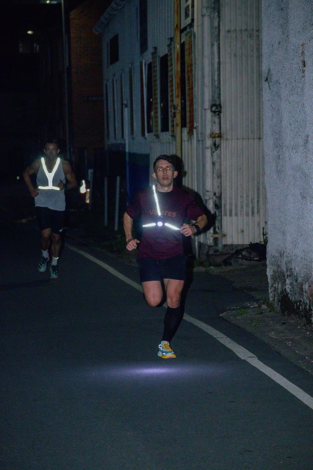 Two runners jogging at night on a dark street, with one runner in the foreground wearing reflective gear and a dark shirt, and another runner in the background wearing a reflective vest.