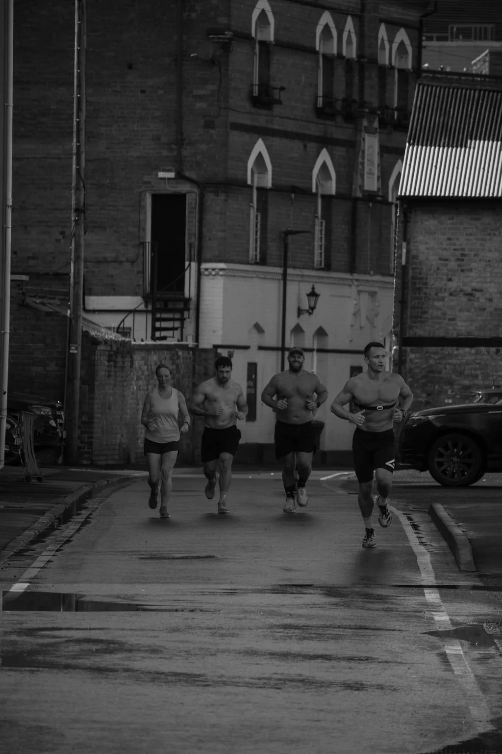 Four people running together on a city street in an urban setting, with brick buildings, a street lamp, and parked cars visible in the background.