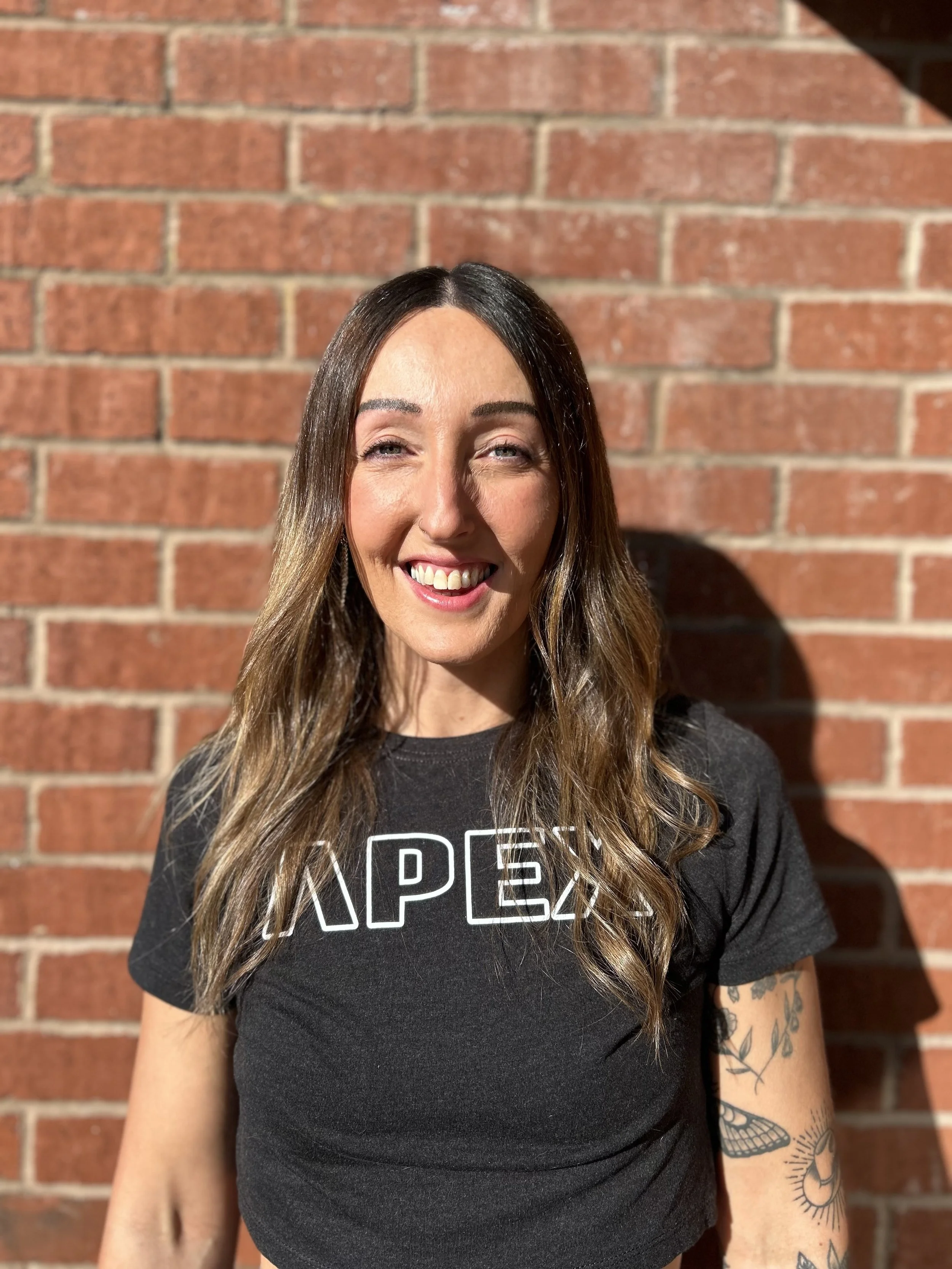 Smiling woman with wavy brown hair standing against a brick wall, wearing a black T-shirt with white text, and tattoos on her left arm.