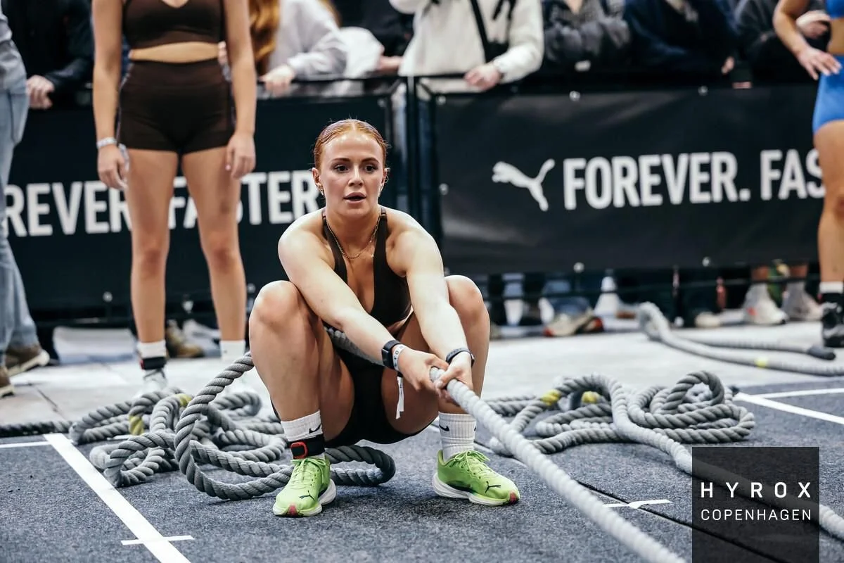 A woman in sportswear is participating in a tug-of-war competition, sitting on the ground while pulling a thick rope. There are spectators and banners in the background, including a Puma logo and text.