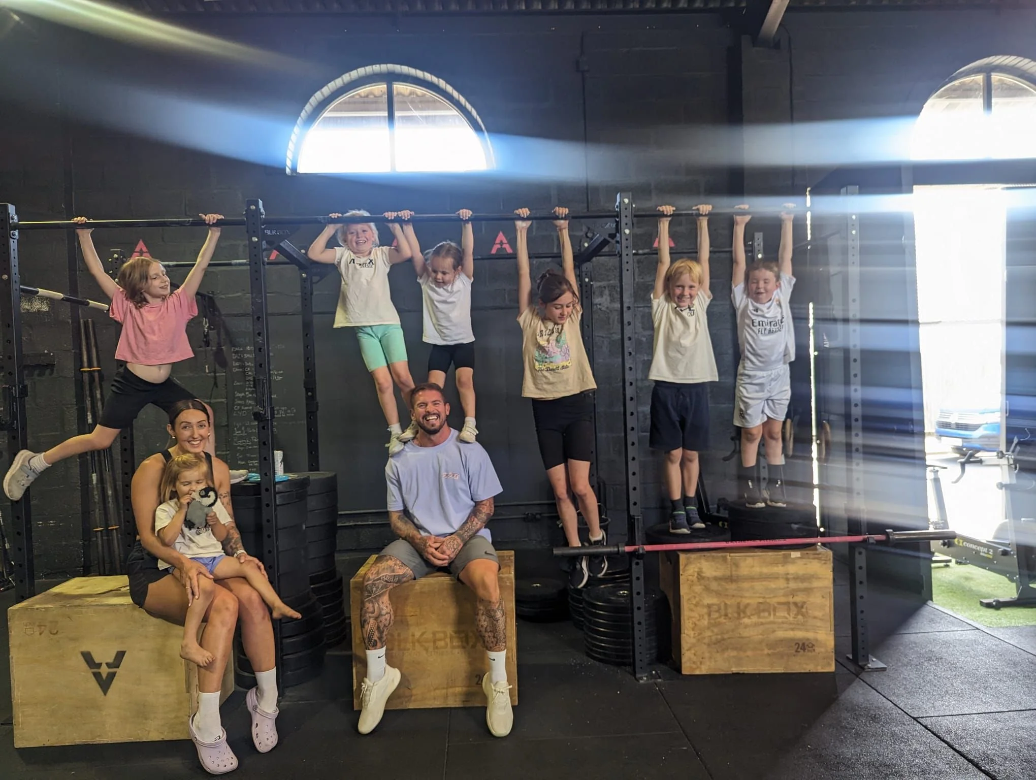 Group of children and two adults at a gym. Some children are hanging from a pull-up bar, while others are standing or sitting on gym boxes and weights. Bright sunlight streaming through windows in the background.