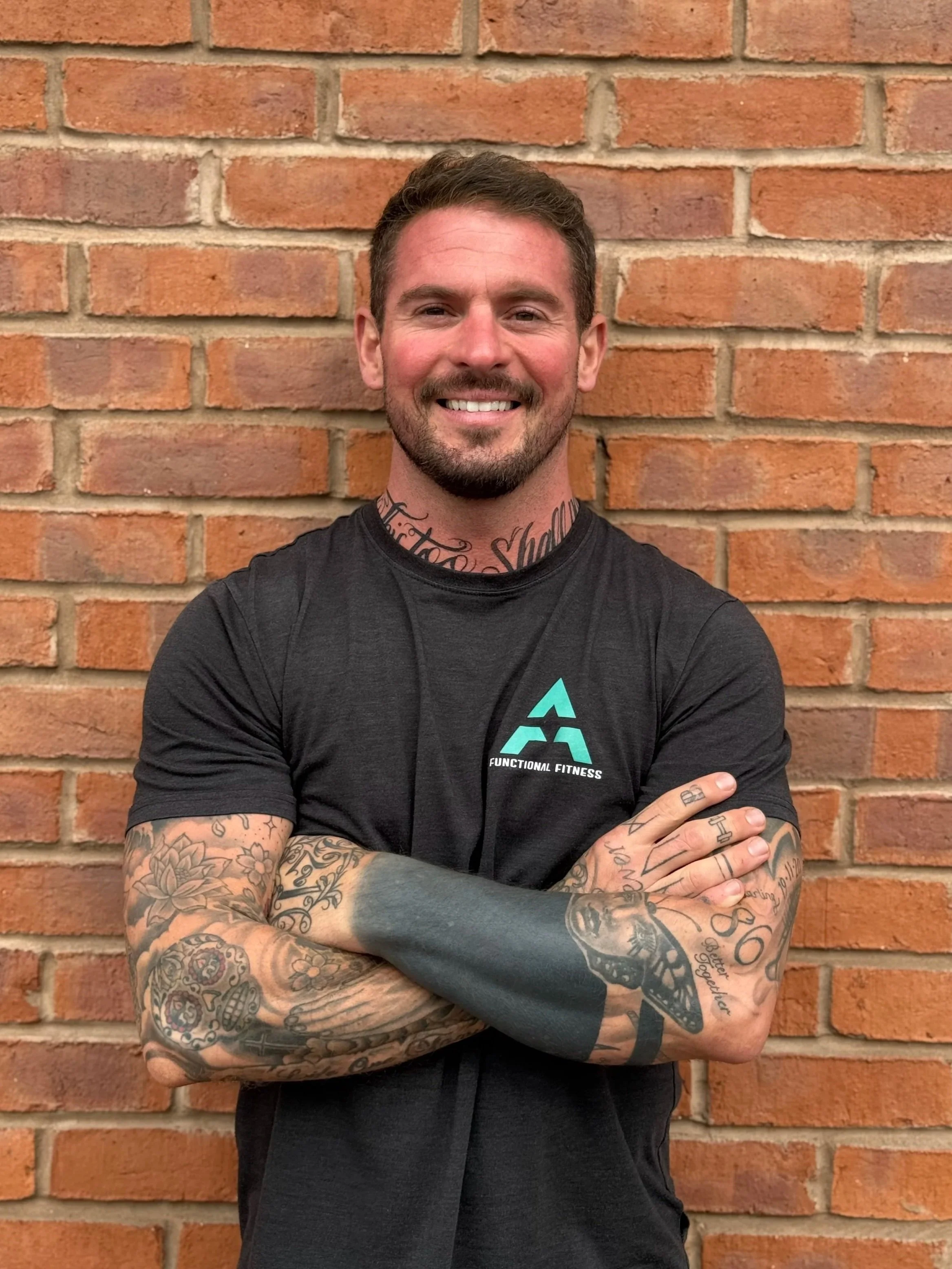 A smiling man with tattoos on his arms and neck, wearing a black athletic shirt with a fitness logo, standing against a brick wall with arms crossed.
