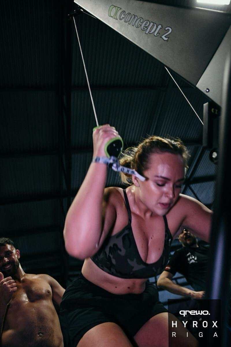 A woman with a gym wristband and camo sports bra exercises on a treadmill, sweating and focused, in a gym with other people. The gym has a dark ceiling with an 'OConcept 2' rower sign visible.