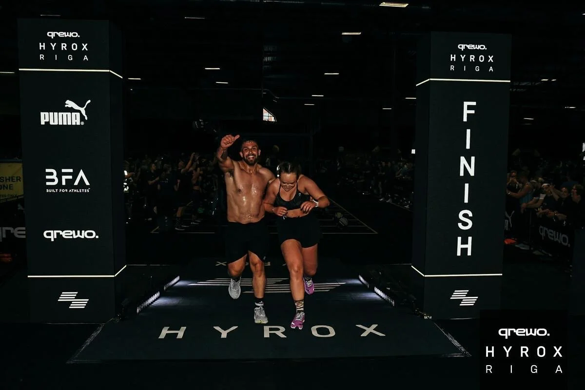 Two people celebrating crossing the finish line of a race at the Hyrox Riga event, with a crowd in the background. One man is shirtless, smiling, and raising his fist, while a woman is dressed in athletic wear, looking down and smiling.