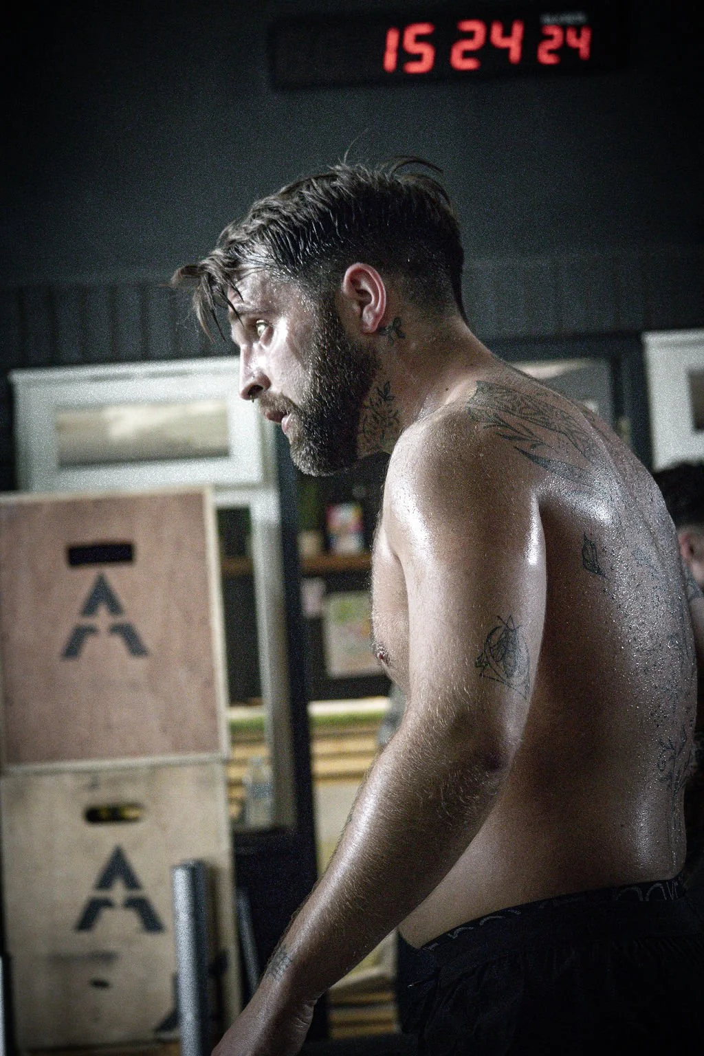 A shirtless man with tattoos and a beard, sweating, inside a gym, with a digital clock showing 15:24:24 in the background.