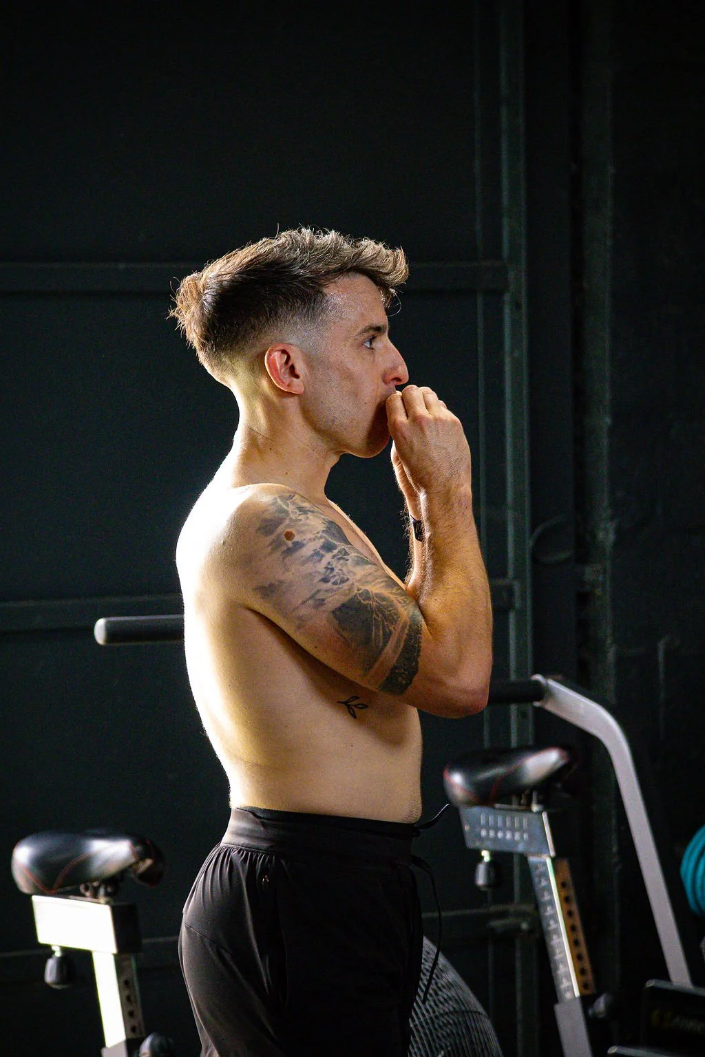 A shirtless man with tattoos on his arm, standing in a gym, appearing to be in deep thought.