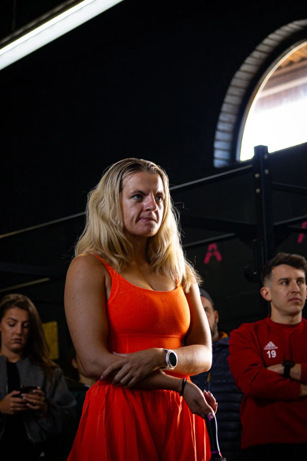 A woman with blonde hair wearing an orange sports top and red skirt stands with arms crossed, looking serious, in a gym or fitness facility with other people in the background.