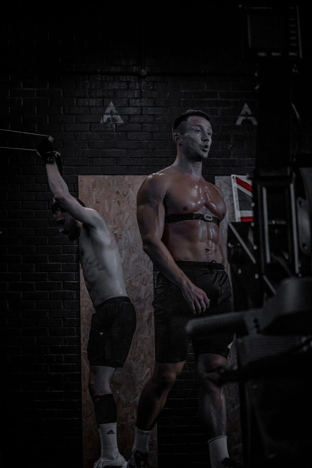 A shirtless man with a body harness is standing in a gym, sweating, with likely a personal trainer or coach nearby, in a dimly lit space with a brick wall background.