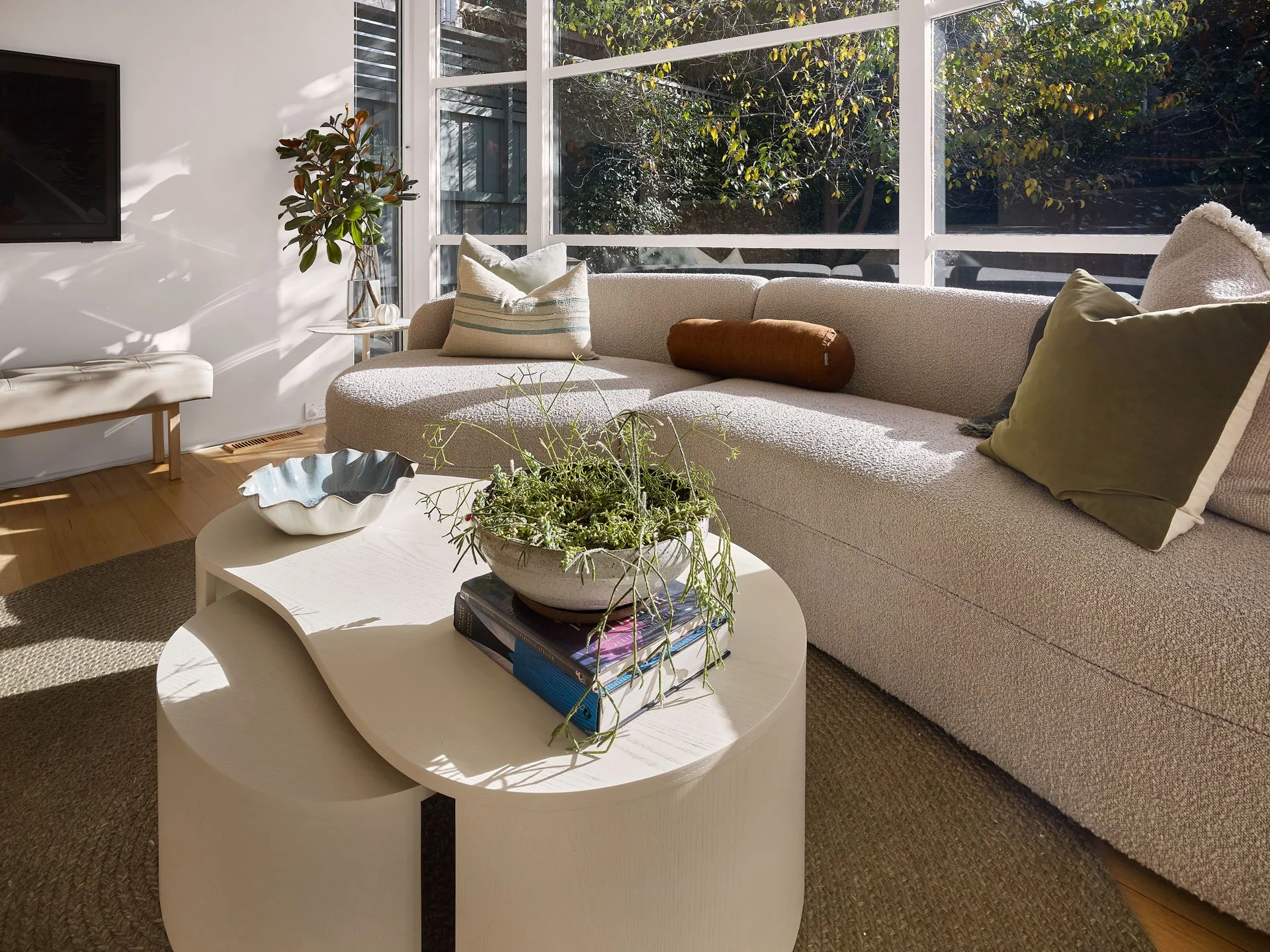 Living room with beige sofa, pillows, plant, and coffee table with books and greenery, large windows with outside trees, sunlight